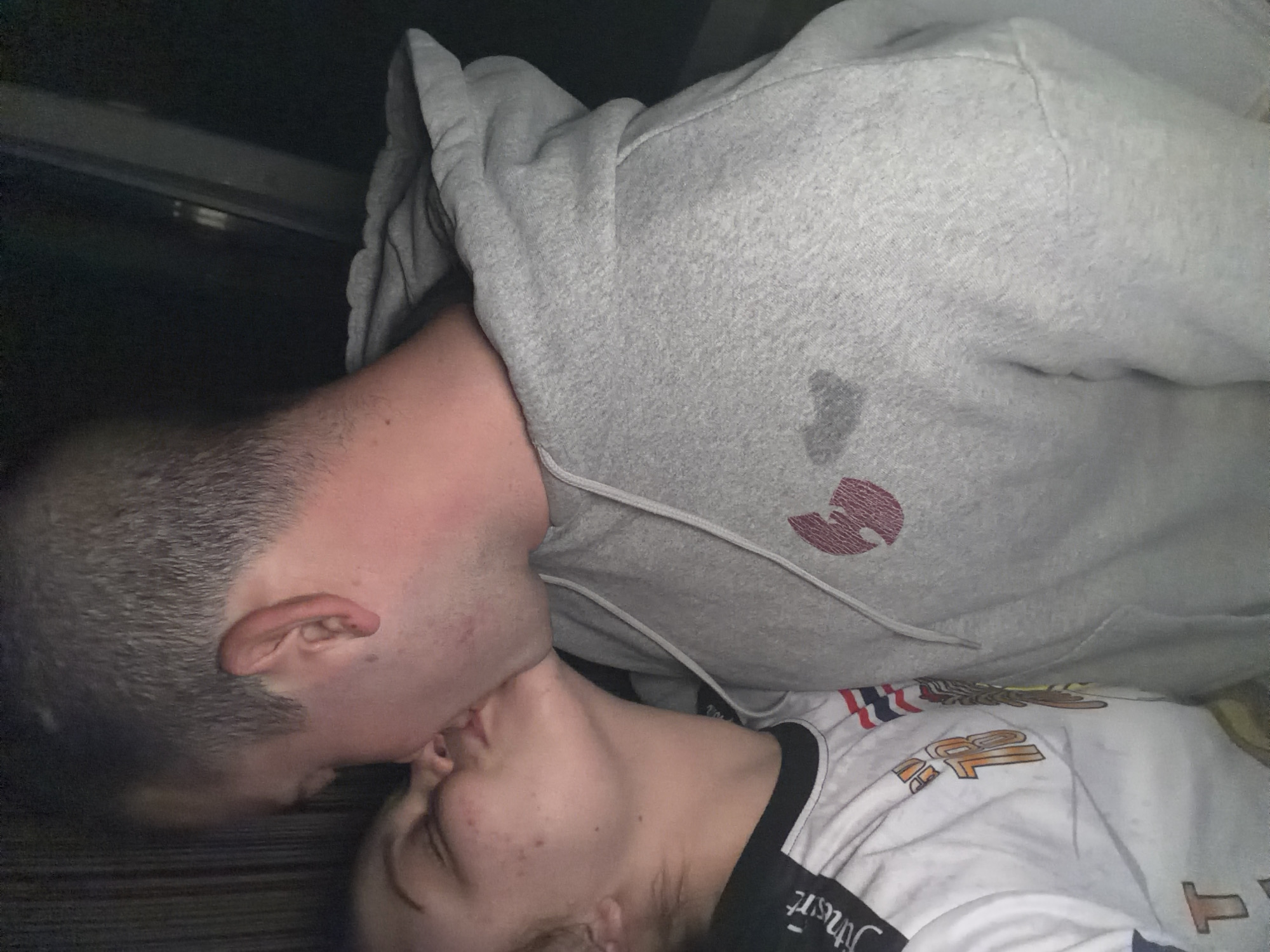 Two people are captured in a close-up, intimate moment, sharing a kiss. The image is vertically oriented, showing their heads and upper torsos. The person on the left has short, dark hair and is wearing a grey hooded sweatshirt. A red Wu-Tang Clan logo is visible on the left chest area of the hoodie, along with a dark mark or shadow above it. Their skin appears fair with a slight redness on the cheek, possibly from the close contact.
The person on the right has lighter hair and is wearing a white t-shirt with a graphic design. Part of an orange "12" is visible on the t-shirt, and there is faint, inverted text on a black trim or collar. This individual also has fair skin, with a few small blemishes visible on their cheek.
The background is dark and indistinct, suggesting a dimly lit indoor setting or a private space, possibly taken at night or in a room with low light. The focus is entirely on the two individuals and their shared moment of affection. The close cropping emphasizes the intimacy of the interaction. This scene takes place in Varna, Bulgaria.