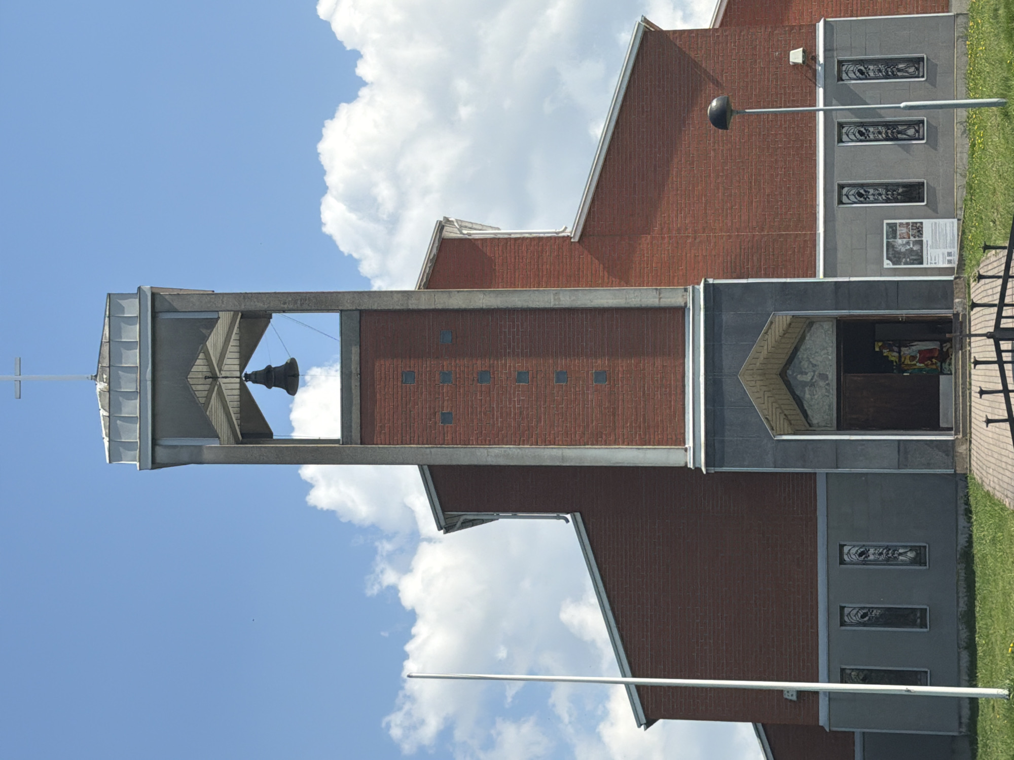 The image shows an exterior view of a building in what appears to be Tokkari, Russia. The architecture features a tall bell tower with a cross at the top, situated next to a brick building. The sky is visible behind the tower, displaying a mix of blue sky and white clouds. To the right of the bell tower, part of the building includes a doorway and stained glass windows. A lamppost is visible near the building's entrance. The area surrounding the building appears to be a grassy lawn or park. There is no readily apparent evidence of people or specific actions.