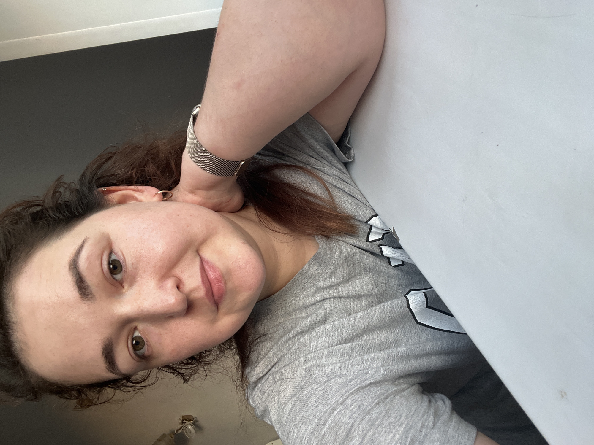 The image shows a young woman, likely in her early to mid-twenties, taking a selfie. She has medium-length, dark brown, curly hair and light brown eyes. She is wearing a grey t-shirt with a graphic print on the front. A silver watch is visible on her left wrist. Her left arm is bent, with her hand resting on the back of her head. She is leaning against a white surface. The background appears to be an indoor setting, possibly a room, with a dark grey wall and a white surface. The lighting suggests it is daytime. The woman has a relaxed and pleasant expression. There is no visible text in the image. The location context is Chelyabinsk, Russia.