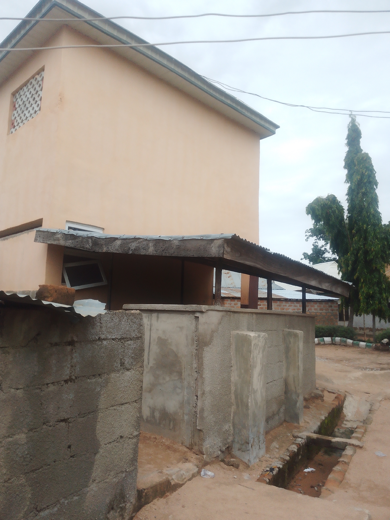 The image shows an outdoor scene in Jalingo, Nigeria, likely during daylight hours. The weather appears overcast, with a cloudy sky dominating the upper portion of the frame. The foreground features a low, rough-hewn concrete wall, part of which is constructed from larger concrete blocks. Adjacent to this wall are two tall, unadorned concrete pillars.
Behind the wall and pillars, a long, low, corrugated metal roof is supported by wooden beams. This roof covers a shaded area with visible brickwork and what appears to be another section of roof behind it. To the left, a portion of a building with light-peach colored walls is visible, featuring a rectangular window with a decorative lattice pattern. Another window, a dark rectangular shape, is tilted and visible within the covered area under the metal roof.
In the background, two tall, slender trees with dense, drooping foliage stand prominently. Their green color contrasts with the muted tones of the structures. A curved border of white and green-striped material, possibly part of landscaping, is visible to the right of the trees. The ground is a mixture of dirt and sand, with some patches of brickwork visible, particularly around a shallow, partially filled drainage channel running along the right side of the image. Some discarded plastic can be seen on the ground. The scene appears to be an exterior of a building complex or compound, possibly a public or institutional space, given the utilitarian nature of the structures. There is no visible text in the image.