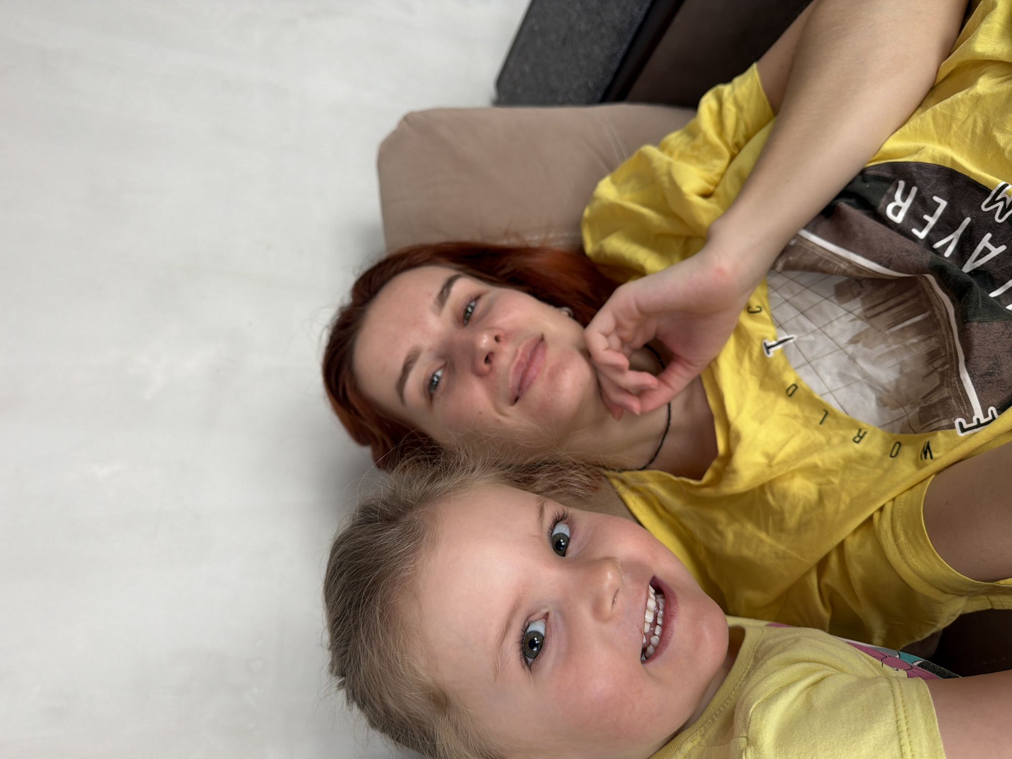 The image shows a woman and a young child lying down together. The woman has red hair and is wearing a yellow shirt with text on it. The child has light hair and is also wearing a yellow shirt. They are smiling at the camera. The background appears to be a light-colored floor. Based on previous knowledge, this image was captured in Chaikovsky, Russia.