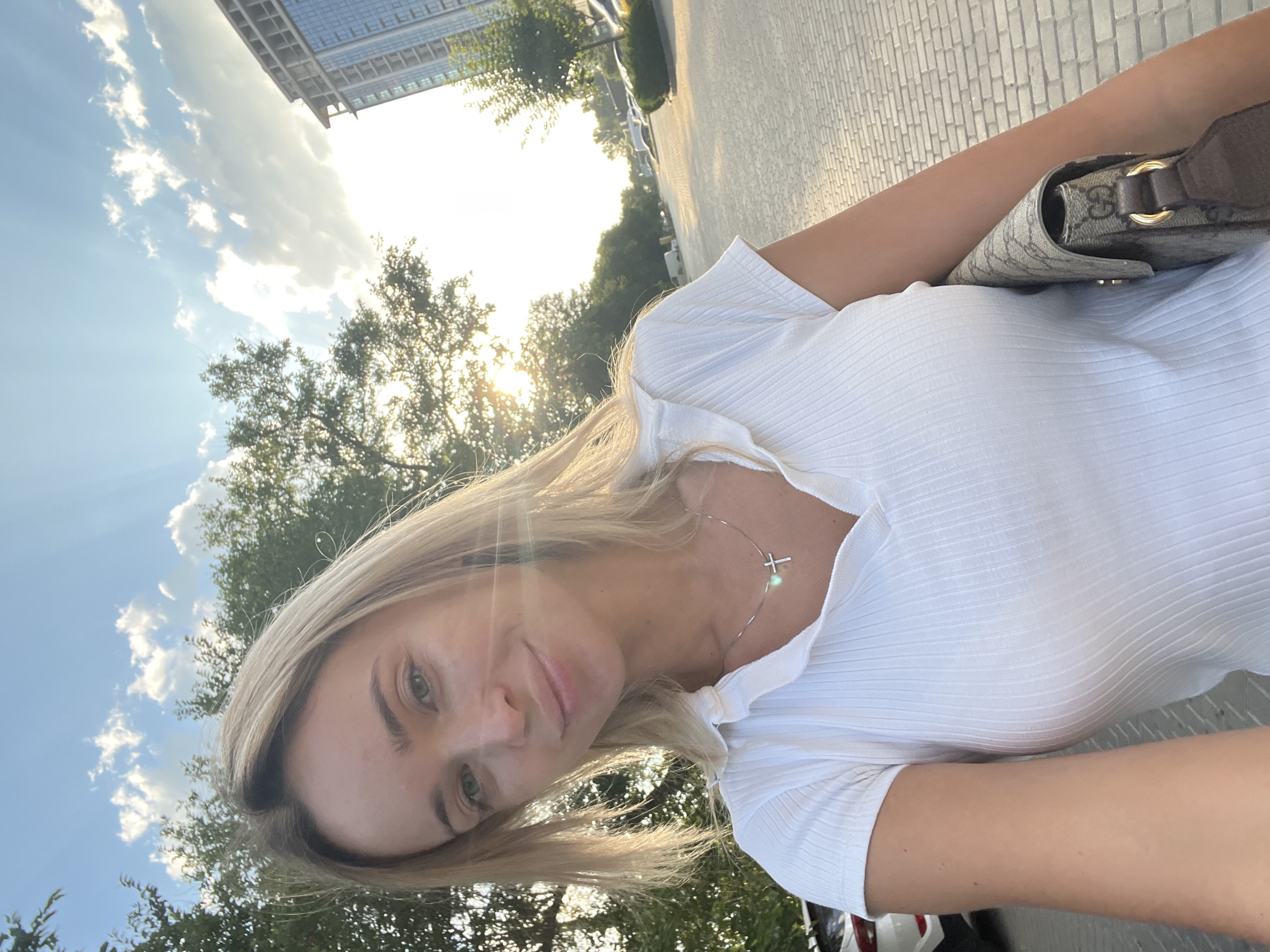 A blonde woman in a white ribbed polo shirt stands on a cobblestone walkway, holding a beige Gucci handbag. Sunlight streams from behind her, creating a lens flare and illuminating the sky with scattered clouds. Trees and a modern building are visible in the background. The location is identified as Fontanka, Ukraine.