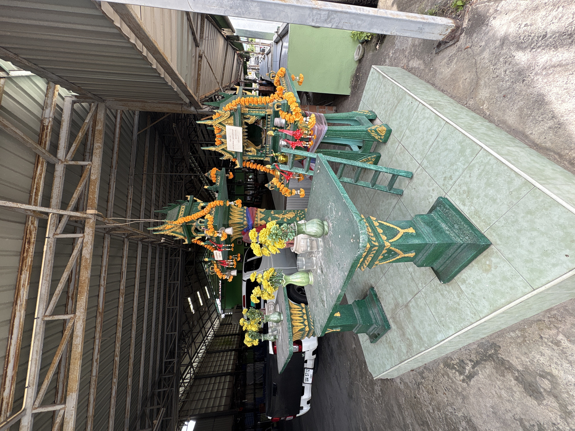In Chalong, Thailand, an outdoor shrine area is depicted under a corrugated metal roof supported by rusting metal beams. The shrine itself is a miniature, ornate structure painted green with gold accents, adorned with yellow marigold garlands and small red tassels. Two green tables with gold detailing sit in front of the shrine on a raised tiled platform. The table on the left holds three green vases with yellow flowers, while the table on the right has two glasses of water and a small green vase with yellow flowers. A small green ladder leans against the shrine structure. In the background, a white pickup truck with a black tarp covering its rear is parked on a concrete floor. Beyond the truck, the interior of a large building with shelving and stacked materials is visible. The scene appears to be an outdoor service area or workshop.