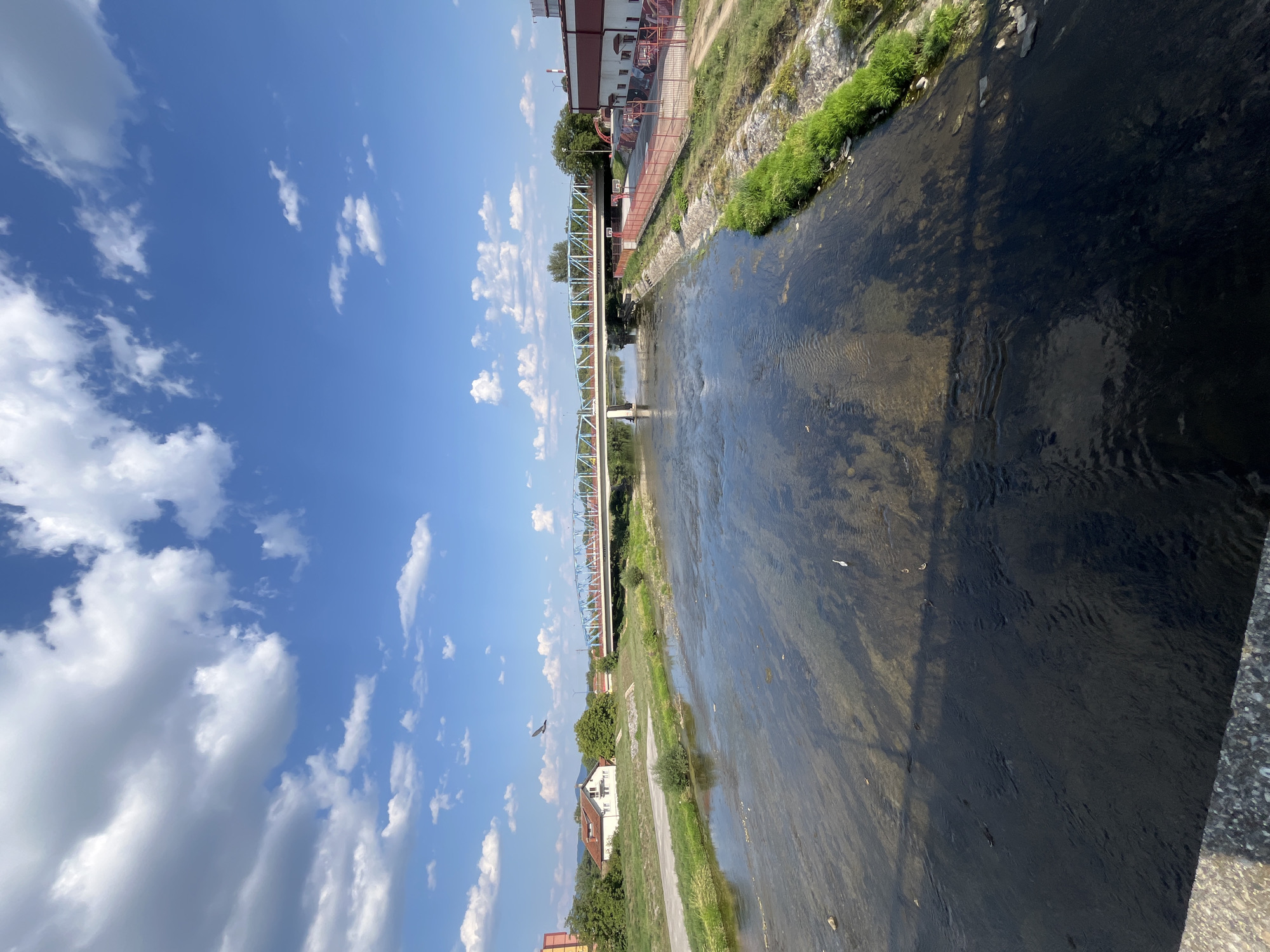 The image depicts a wide, shallow river flowing through an urban area, likely in the city of Čačak, Serbia. A blue and white truss bridge spans the river, with a road running across it. On the right bank, a modern building with red and white accents is visible, adjacent to a fenced outdoor sports court. The left bank features a grassy area with a white house and trees. The riverbed is visible through the clear water, with some rocks and vegetation. The sky is blue with scattered white clouds, and a bird is in flight overhead. The overall scene suggests a sunny day with moderate urban development.
