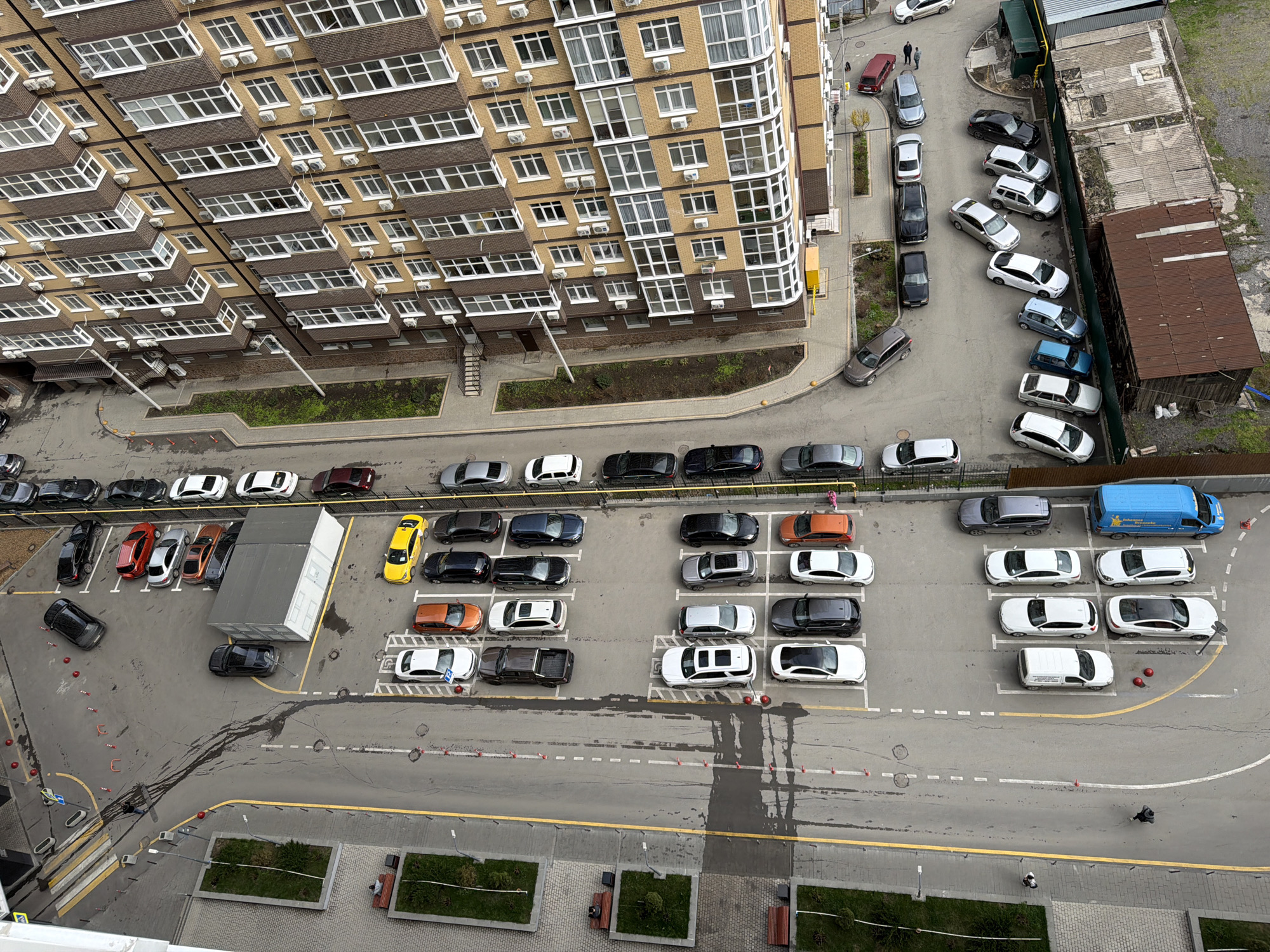 The image presents an aerial view of a residential area, specifically a parking lot next to a multi-story apartment building.
**Location:** The image likely originates in Azov, Russia, although this cannot be definitively confirmed from the image alone.
**Environment:** The environment appears to be urban, with a large apartment building taking up a significant portion of the frame. There's a parking lot adjacent to the building, with several rows of parked cars. Vegetation is visible in the form of planted beds and some grassy areas near the building.
**People:** There are a few individuals visible, including one walking in the parking area and others near the top right corner of the frame.
**Objects:**
* **Building:** A large, multi-story apartment building with light-colored brick and numerous windows.
* **Vehicles:** A large number of cars are parked in an organized parking lot.
* **Parking Lot:** Marked parking spaces and a road with a yellow line. There are also small traffic cones or markers visible.
* **Landscaping:** Planted beds with greenery are spaced out near the bottom of the frame.
* **Miscellaneous:** Possible temporary structure in the center of the parking lot.
**Actions:** People are walking in the vicinity of the building and parking lot. Cars are parked.
**Interactions:** The interaction is mainly between people and their environment (walking, parking). The vehicles are parked in their designated spots. The architectural elements are used for housing and parking.