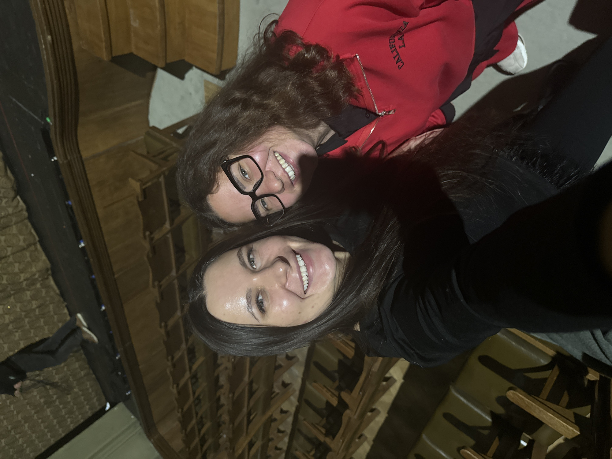 Two young women are posing for a selfie in what appears to be a church interior. The woman in the foreground has dark hair and is wearing a black top. The woman behind her has curly hair and is wearing glasses and a red jacket. The background consists of rows of wooden pews. Based on the architectural style and the presence of pews, the location is likely a Russian Orthodox church in Moscow. The image appears to be taken inside the church due to the lighting and architecture.