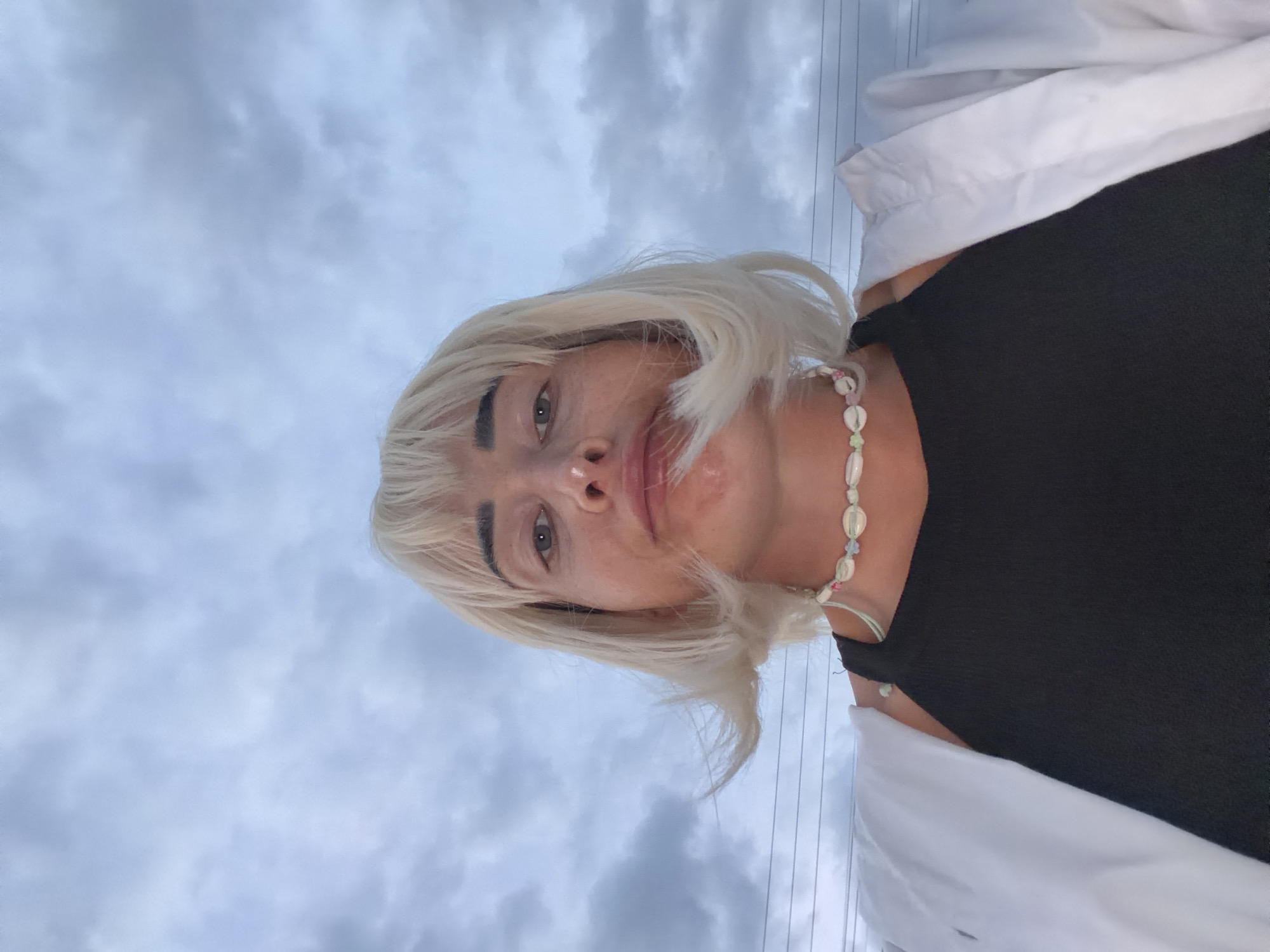 A person with blonde hair and a black tank top is shown against a cloudy sky. They are wearing a necklace made of seashells. The photo appears to be a selfie taken outdoors. The clouds are dense and dark, suggesting overcast weather. The person's face is partially obscured by their hair, which is being blown by the wind. The overall impression is casual and informal. There is no indication of the location being Sochi, Russia.