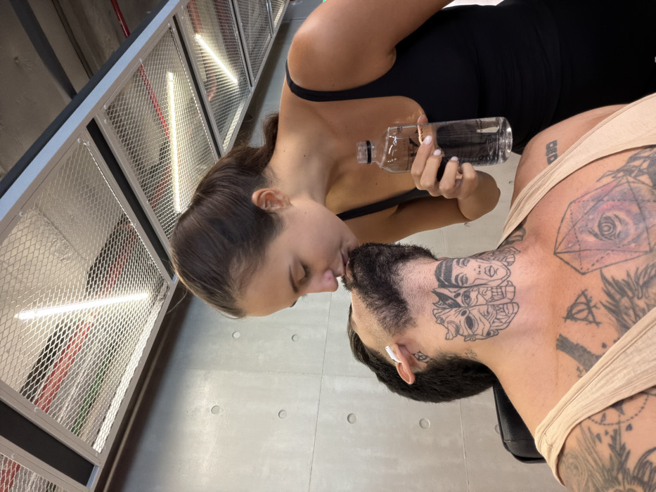 This image, captured indoors in Bangkok, Thailand, displays two individuals. A female, with dark hair pulled back in a ponytail and wearing a black tank top, is shown leaning in to kiss the chin/beard area of a male. Her eyes are closed, and she holds a clear plastic water bottle in her right hand. The male, wearing a light-colored tank top, has a dark beard, a white earbud in his left ear, and multiple tattoos. Prominent tattoos on his neck depict three distinct faces, while his chest features a large tattoo of a geometric crystal with an eye inside, alongside other designs. The background consists of a light grey concrete wall with visible circular imprints and an industrial-style ceiling with metallic mesh panels, linear fluorescent light fixtures, and partially visible red piping. The scene is brightly illuminated by artificial overhead lighting.