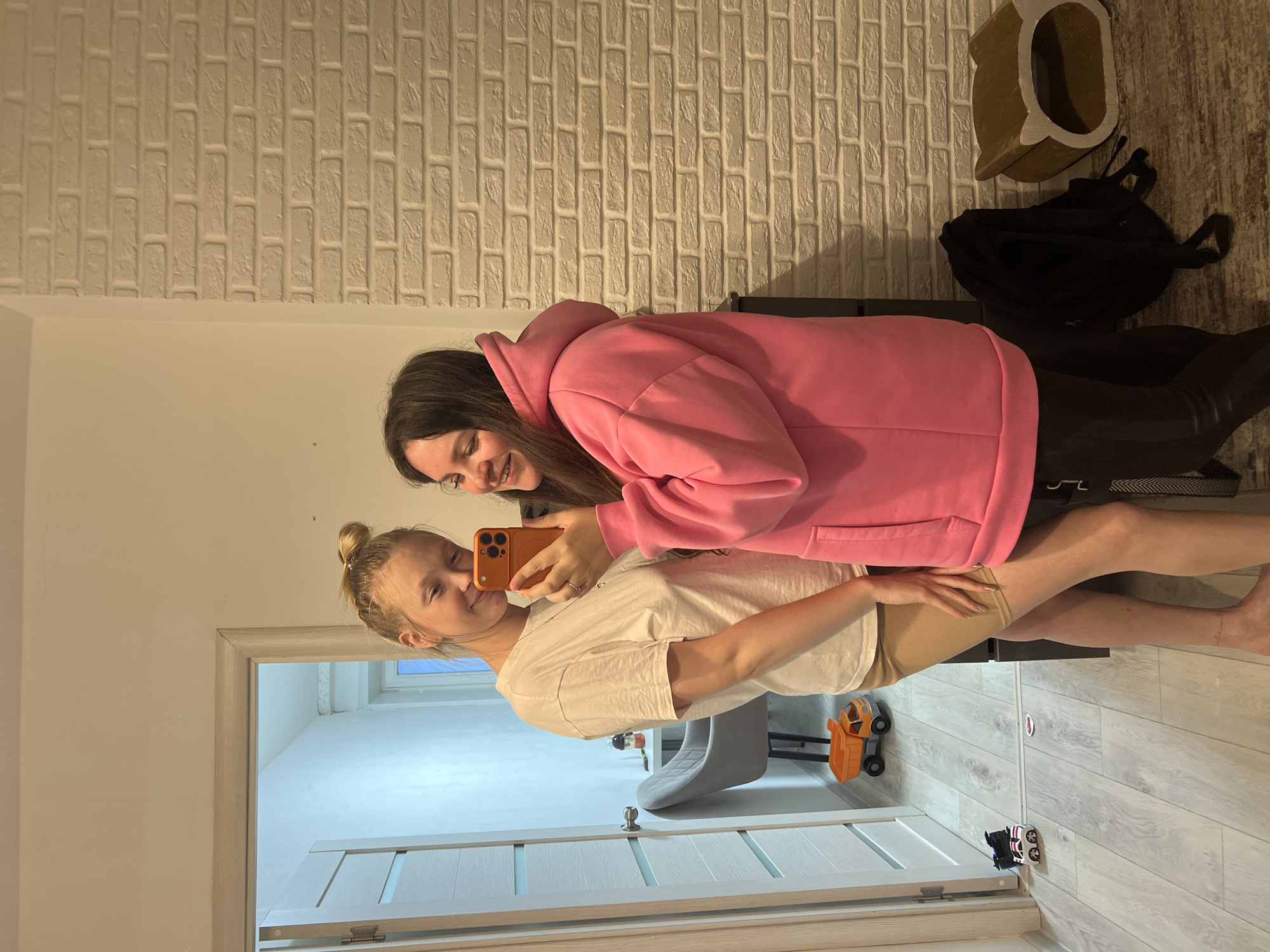This image captures an indoor scene in the city of Chaikovsky, Russia, featuring two young adult women taking a selfie.
The woman on the left has blonde hair styled in a bun, a fair complexion, and is wearing a white short-sleeved t-shirt and light brown or tan cycling shorts. She is barefoot, has an anklet on her left ankle, and is smiling while looking towards the phone.
The woman on the right, actively taking the selfie, has long dark hair and is wearing an oversized bright pink hooded sweatshirt and dark trousers. She is holding an orange-cased smartphone (with a visible triple-camera array) in her right hand, positioned to capture the image, and is also smiling towards the phone.
The background consists of two distinct wall textures: a plain light-colored painted wall on the left side of the frame and a white or light beige brick-patterned wall on the right. The floor is light-toned laminate or wood-effect, with a light-colored transition strip running across it.
On the left side, a white door frame leads into another room. Through the slightly ajar door, a grey upholstered chair, a small table with a water bottle, and a window providing natural light are visible. On the floor in the foreground, near the door, are an orange toy dump truck and two smaller toy cars (one black, one white with red stripes).
On the right side, a dark cabinet or piece of furniture is partially visible behind the woman in pink. On the floor next to her are a black backpack and a brown, semi-cylindrical object with an oval opening, resembling a pet bed.