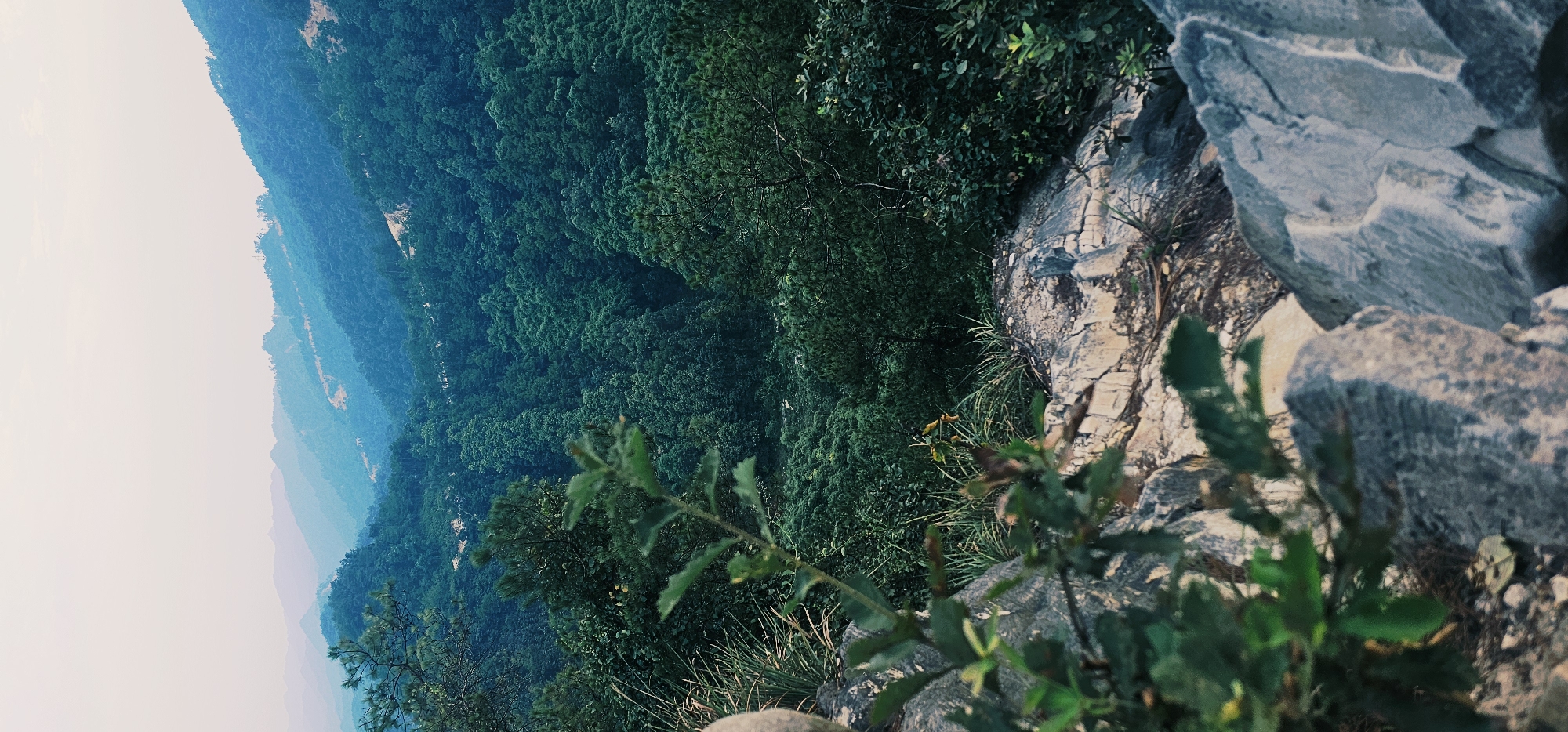 A vertical shot depicts a scenic view of lush, verdant mountains fading into a hazy, pastel sky. In the foreground, weathered grey rocks and a few green leaves frame the scene. The mountains, densely covered in trees, recede in layers, with the distant peaks appearing in shades of blue and purple due to atmospheric perspective. A narrow, winding road is faintly visible on one of the slopes. The overall impression is one of natural beauty and tranquility, characteristic of the mountainous terrain in the Chongqing High-tech Zone, China.