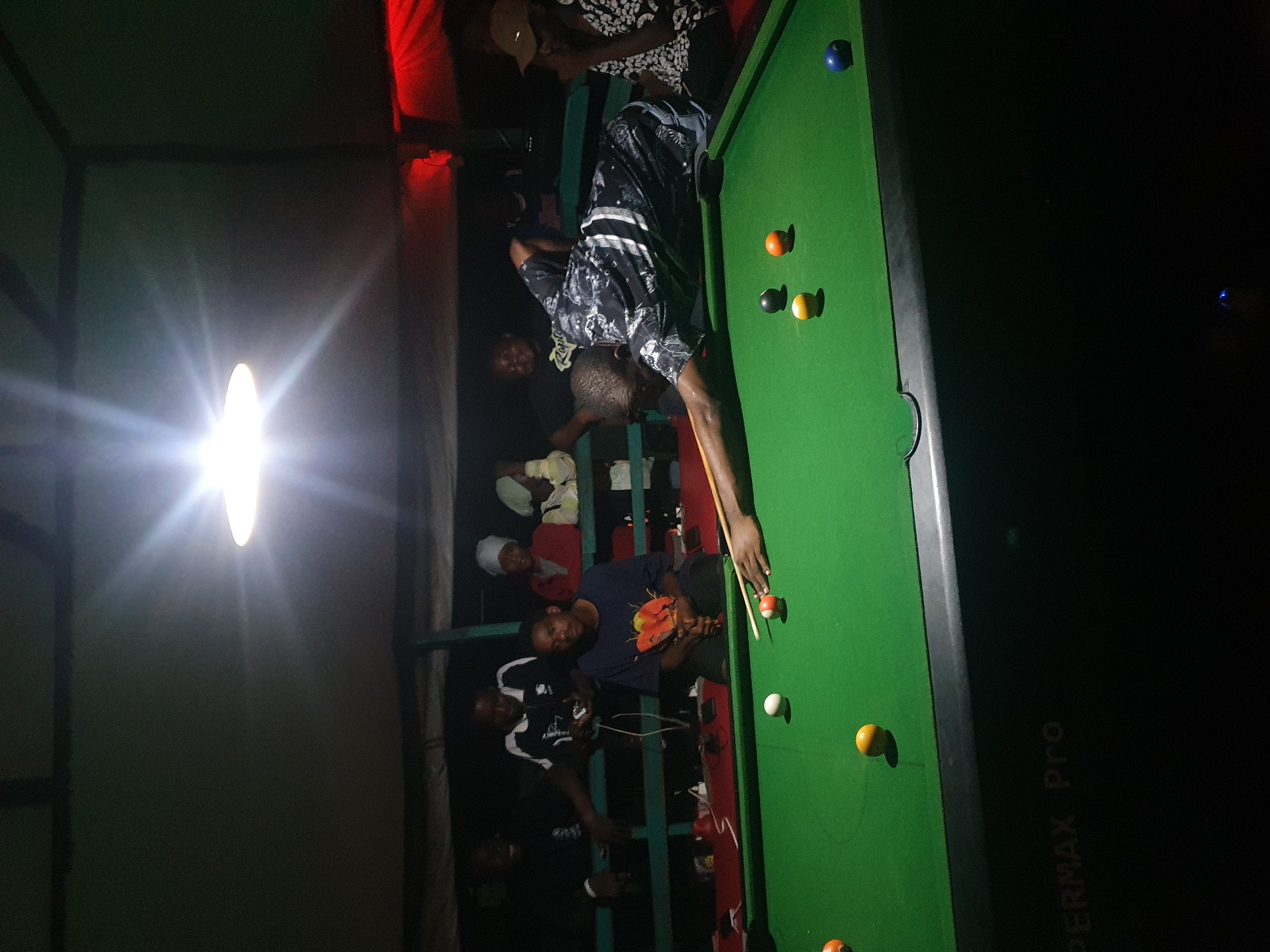 A group of men are gathered around a pool table in Lokoja, Nigeria, under a bright overhead light. One man is actively playing, bending over the table to take a shot with a cue stick, while several others watch from behind. The pool table is green with various colored balls scattered across its surface, including a white cue ball. The environment appears to be a casual, possibly outdoor or semi-outdoor setting, indicated by the visible ceiling structure and the dim lighting in the background. The men are dressed in casual attire. Some are seated on benches or a raised platform behind the pool table. The overall scene depicts a social gathering centered around a game of billiards.