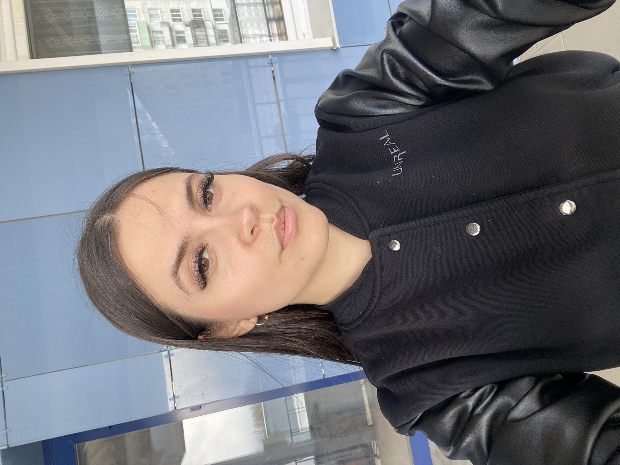 The original image is oriented 90 degrees counter-clockwise, depicting a young woman posing for a photo, likely in an urban or suburban environment during the daytime, potentially in Chaikovsky, Russia.
The subject is a woman with long, dark brown hair and brown eyes. She wears light makeup, including prominent, long false eyelashes and a subtle lip color. Her expression is calm and neutral as she looks towards the camera.
She is dressed in a black jacket featuring contrasting black faux leather sleeves and a ribbed collar. On the left side of her jacket, the word "UNREAL" is embroidered vertically in a light, possibly white or silver, thread. Several shiny metal snap buttons are visible along the front opening of the jacket. A small gold hoop earring is visible in her right ear.
The background consists of a large, light blue, paneled surface, which appears to be a building facade or wall. Above this blue surface, a window reflects parts of other buildings, revealing multiple windows and structural elements, hinting at an architectural setting. The bright and even lighting suggests it is daytime.