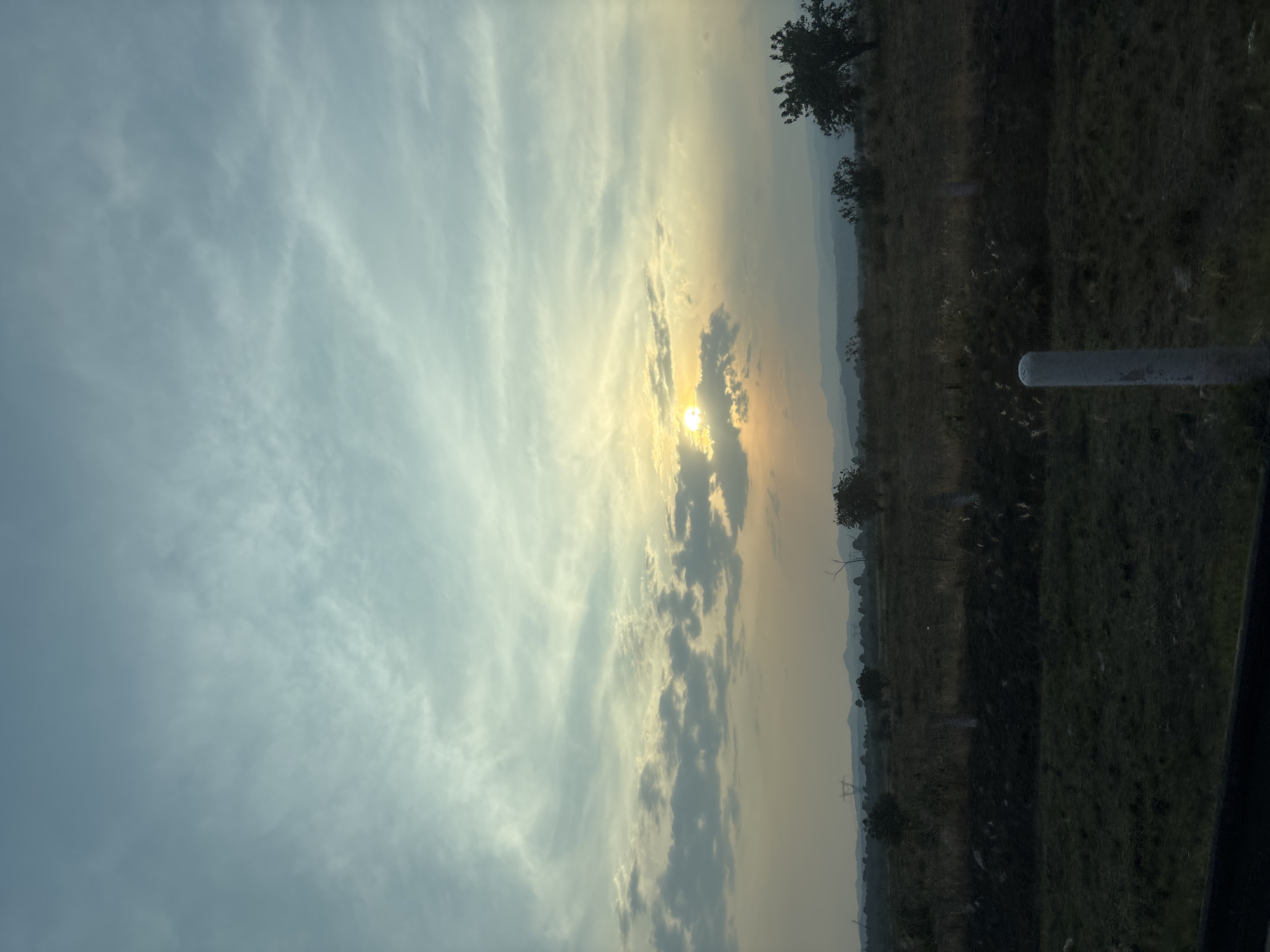 The image shows a landscape at sunset in an undetermined location. The sky, which dominates the upper left portion of the image, features a mix of light blue and gray clouds, with the setting sun visible through a gap in the cloud cover. The horizon line is visible in the lower right of the image, with a stretch of what appears to be a field or open space. The land is uneven and partially obscured by darkness, suggesting the time of day is late afternoon or dusk. Silhouettes of trees are visible along the horizon line. A cylindrical object (possibly a pole or railing) is visible in the lower right corner. The image lacks specific details needed to conclusively determine the location or specific details about the environment. No people or interactions are visible. The setting is rural or suburban. There is no information to conclude that this is located in Metepec, Mexico.