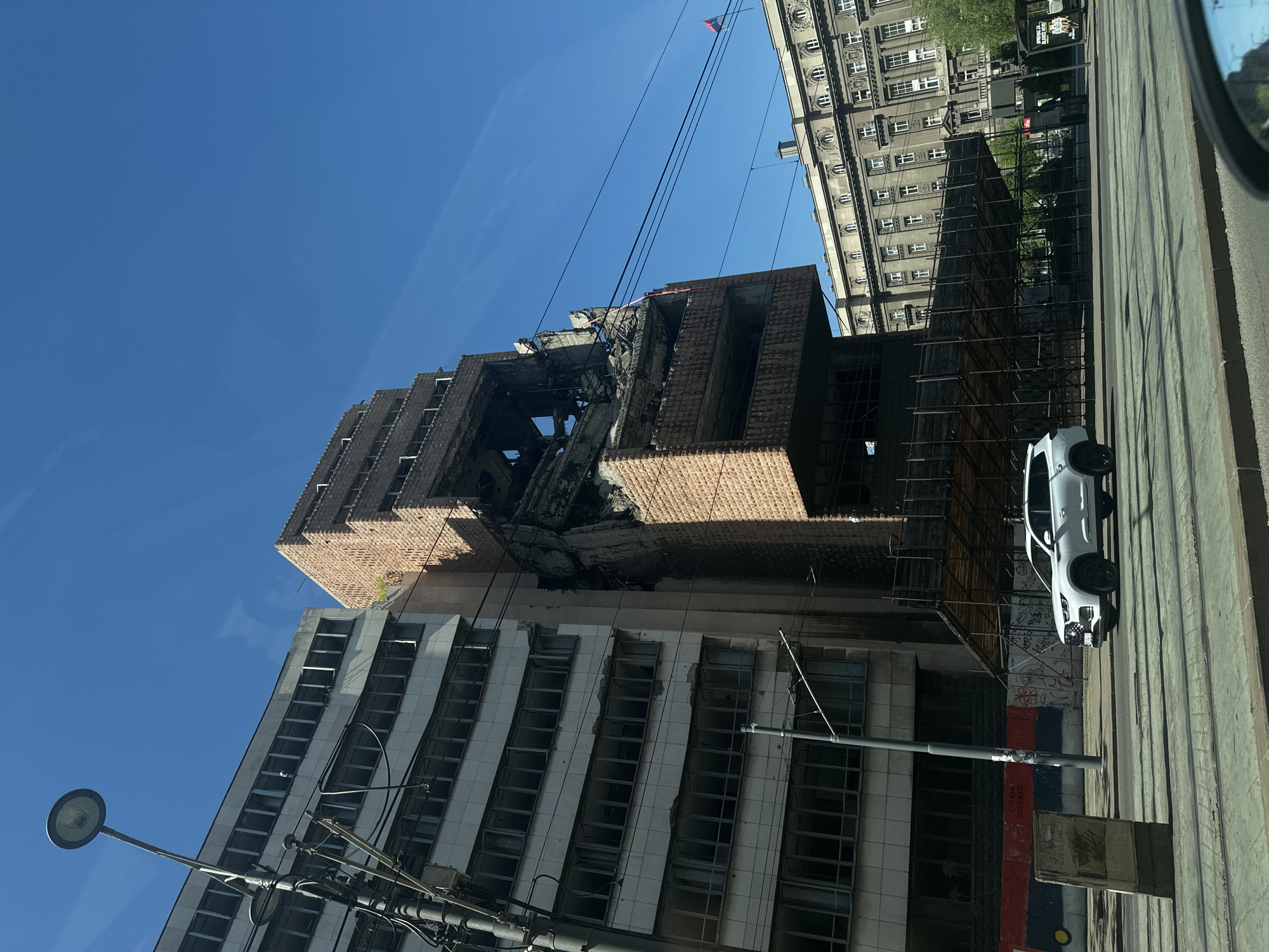 The image depicts the bombed-out shell of the Yugoslav Ministry of Defense in Belgrade, Serbia, with clear blue skies above. The building is partially demolished, showing exposed brick and concrete. Scaffolding supports a section of the damaged structure. A modern white car is parked on the street next to the building, indicating the presence of potential passersby or onlookers. A neighboring building appears in the background, displaying a stark contrast between past conflict and contemporary urban life. A streetlight and various utility lines are visible in the foreground, adding to the urban environment. The presence of graffiti on the remaining walls indicates the building's status as an abandoned or neglected site.