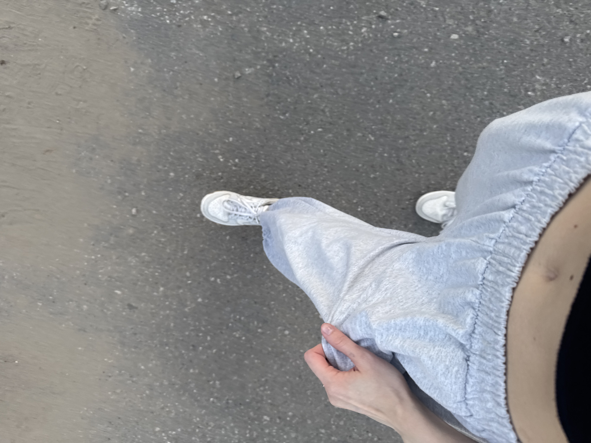 The image shows a top-down view of a person's legs and torso, standing on a grey asphalt surface. The person is wearing light grey sweatpants and white sneakers. A hand is resting on the left thigh. The background is an out-of-focus view of the asphalt. There is no identifiable information within the image to suggest a specific location in the city of Perm, Russia.