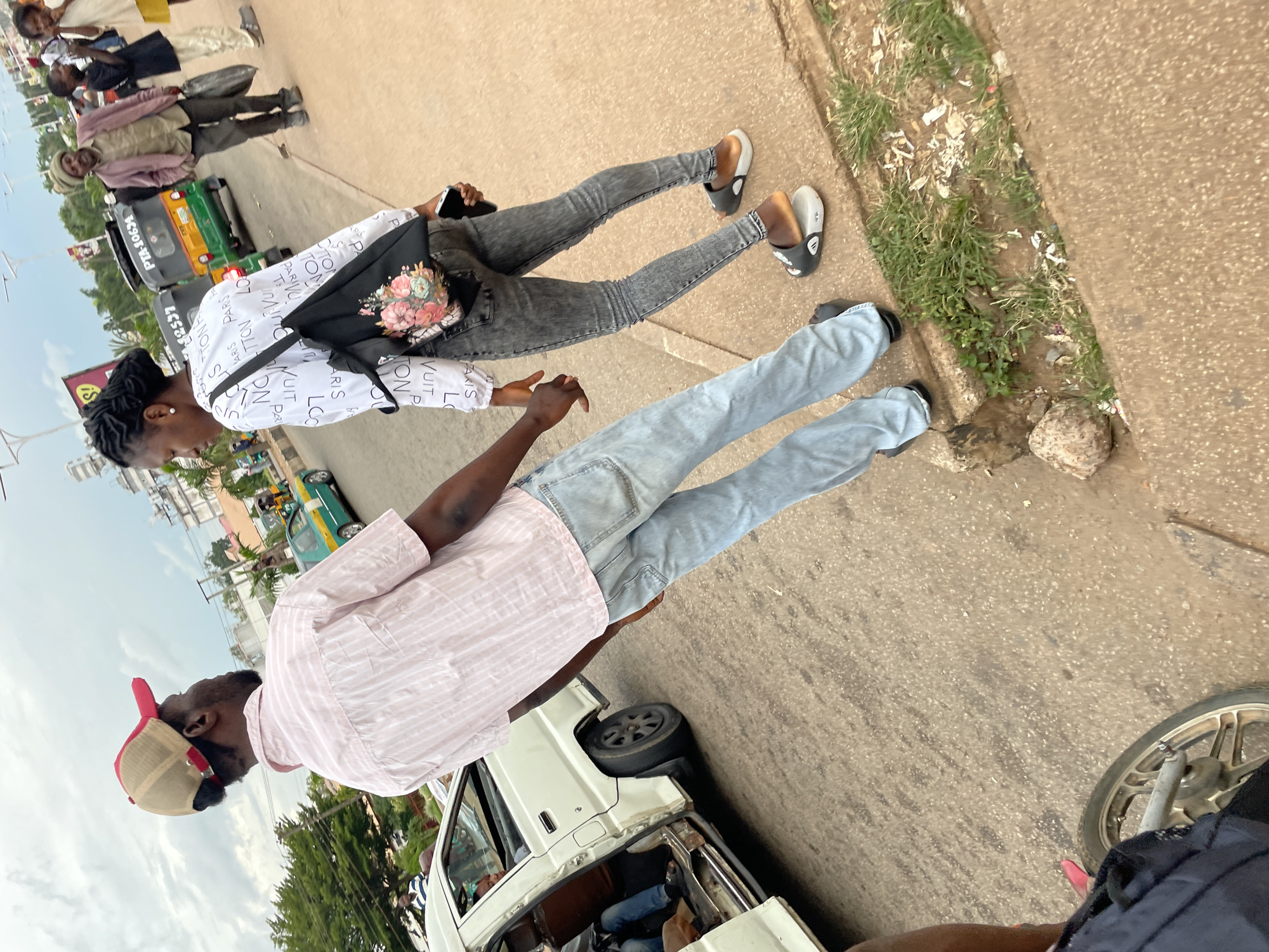 In Jos, Nigeria, a man in a pink striped shirt and a red and tan baseball cap stands with his back to the camera, facing a woman wearing a white top with black lettering and gray jeans. The woman has her hair in braided extensions and is carrying a black floral-patterned bag over her shoulder and holding a smartphone in her right hand. They are on a paved street, with a white passenger van to their left and a green and yellow auto-rickshaw in the background. Other people are visible on the sidewalk and in the background, along with buildings and trees. The scene appears to be a street in a commercial or residential area of the city.