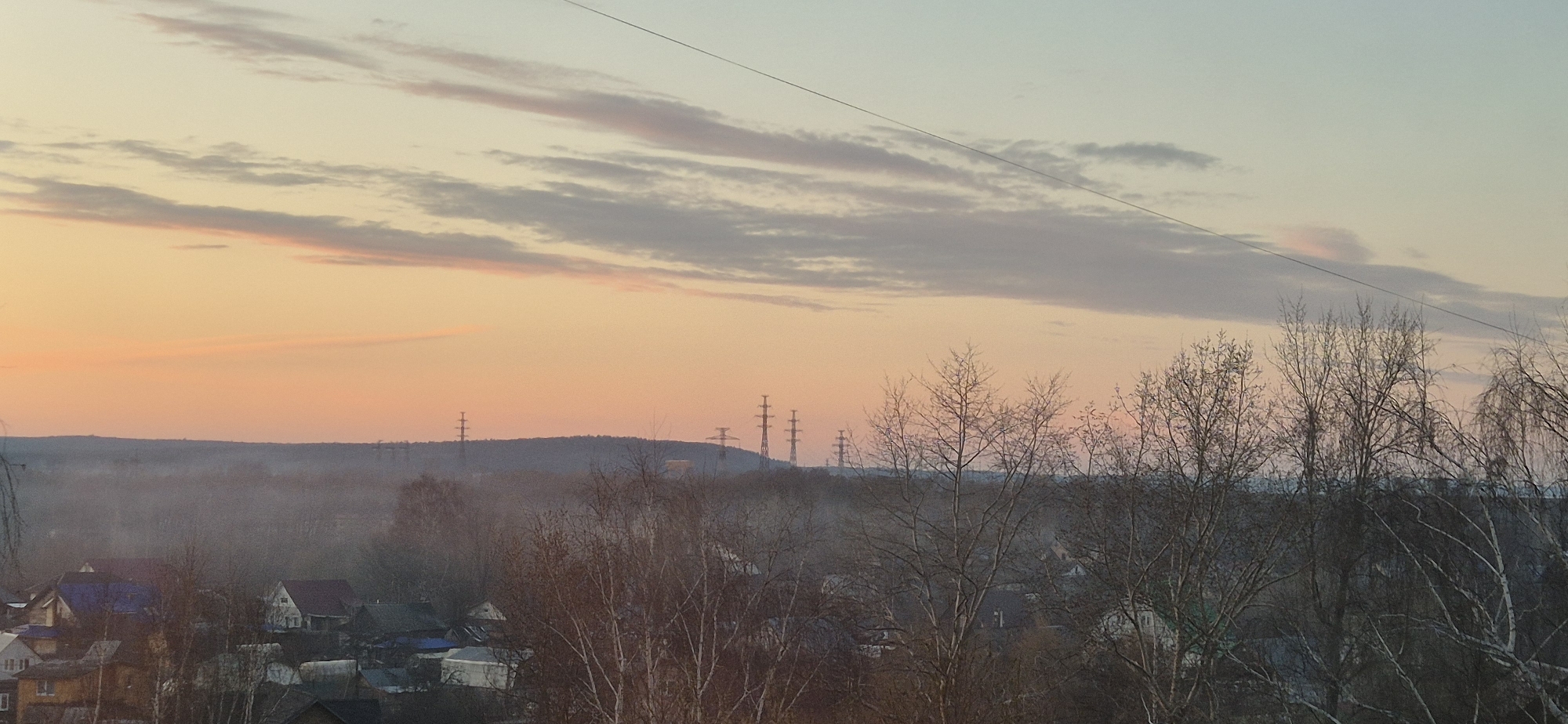 This image captures a tranquil landscape scene at what appears to be either sunrise or sunset, given the soft, warm hues in the sky. The upper portion of the frame is dominated by a gradient sky, transitioning from a warm, golden orange near the horizon to a lighter, pale blue-grey higher up. Scattered across the sky are elongated clouds, some tinged with subtle pinks and purples, running horizontally. A thin, dark wire or power line stretches diagonally from the upper left to the upper right of the frame.
In the mid-ground, a dark, undulating line of hills or distant terrain forms the horizon, behind which the warm glow of the sky is most intense. Silhouetted against this glow are several tall, metal transmission towers or power pylons, spaced out along the ridgeline of the hills. A soft, light haze or fog blankets the valley area between the foreground and the distant hills, obscuring some details and adding depth to the scene.
The foreground is filled with numerous bare-branched trees, their intricate, dark silhouettes creating a natural frame. Their leafless state suggests either late autumn, winter, or early spring. Among these trees, particularly in the lower left, a cluster of residential houses is visible. These houses feature varied roof colors, including dark blue, brown, and light grey, some of which might be lightly dusted with snow or frost. There are no people visible, and no specific activity is taking place; the scene is static and serene. There is no visible text in the image. The overall impression is one of quiet contemplation in a semi-rural or suburban setting during twilight hours.