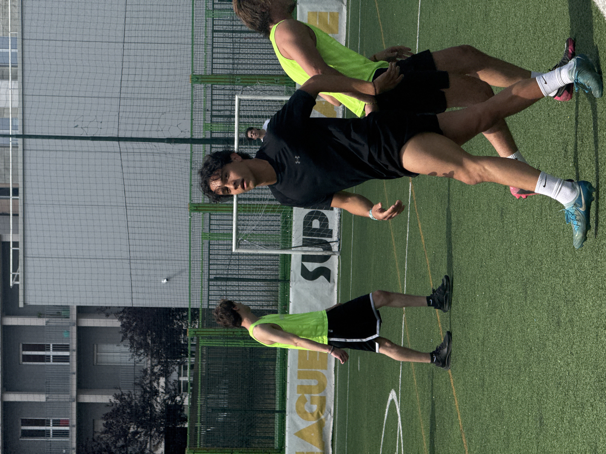 The scene depicts an outdoor artificial turf soccer field under bright daylight in the city of Milan, Italy. Four male individuals are visible on the field.
In the foreground, a male with dark, curly hair is positioned slightly left of center, walking towards the right. He wears a black short-sleeved Under Armour t-shirt, black athletic shorts, white socks, and blue Nike Tiempo soccer cleats with white laces. A dark, irregular mark is present on his upper left thigh. He looks towards the right. Immediately to his right and slightly behind, a second male is visible, facing towards the right. He wears a neon yellow sleeveless tank top, black shorts with a white side stripe, white socks, and blue soccer cleats (one with a pink sole and black upper, the other blue/green). His light brown hair is disheveled.
Further back on the left, a third male, also in a neon yellow sleeveless tank top and black shorts with a white side stripe, black socks, and black athletic shoes, walks towards the right. His brown hair is short and wavy. In the deep midground, centered behind the main figures and near a white soccer goal, a fourth male in a white t-shirt is partially visible, facing forward.
The soccer field displays white and yellow painted lines. Behind the field, a green metal fence with mesh extends horizontally. Beyond this fence, a partially visible white and yellow banner with black lettering spells "AGUE" and "SUP". Dark green foliage and trees are present behind the banner and fence. In the background, a multi-story grey building with balconies and white-framed windows is visible on the left, while another large grey building structure with a mesh net in front of it stands on the right. A tall green pole rises in front of the right-hand building.