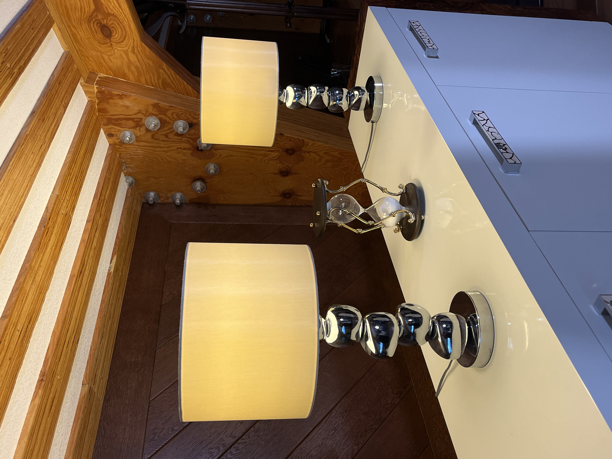 Two table lamps with cream-colored shades sit on a white cabinet. The lamp on the left has a stacked, spherical chrome base, and the lamp on the right has a stacked, cylindrical chrome base. Between the lamps is a small hourglass with a dark base and frame, containing white sand. The cabinet has two doors with decorative chrome handles. The background features dark wood paneling and a wooden ceiling with exposed beams and white panels. The scene appears to be indoors. There are no people or animals present.