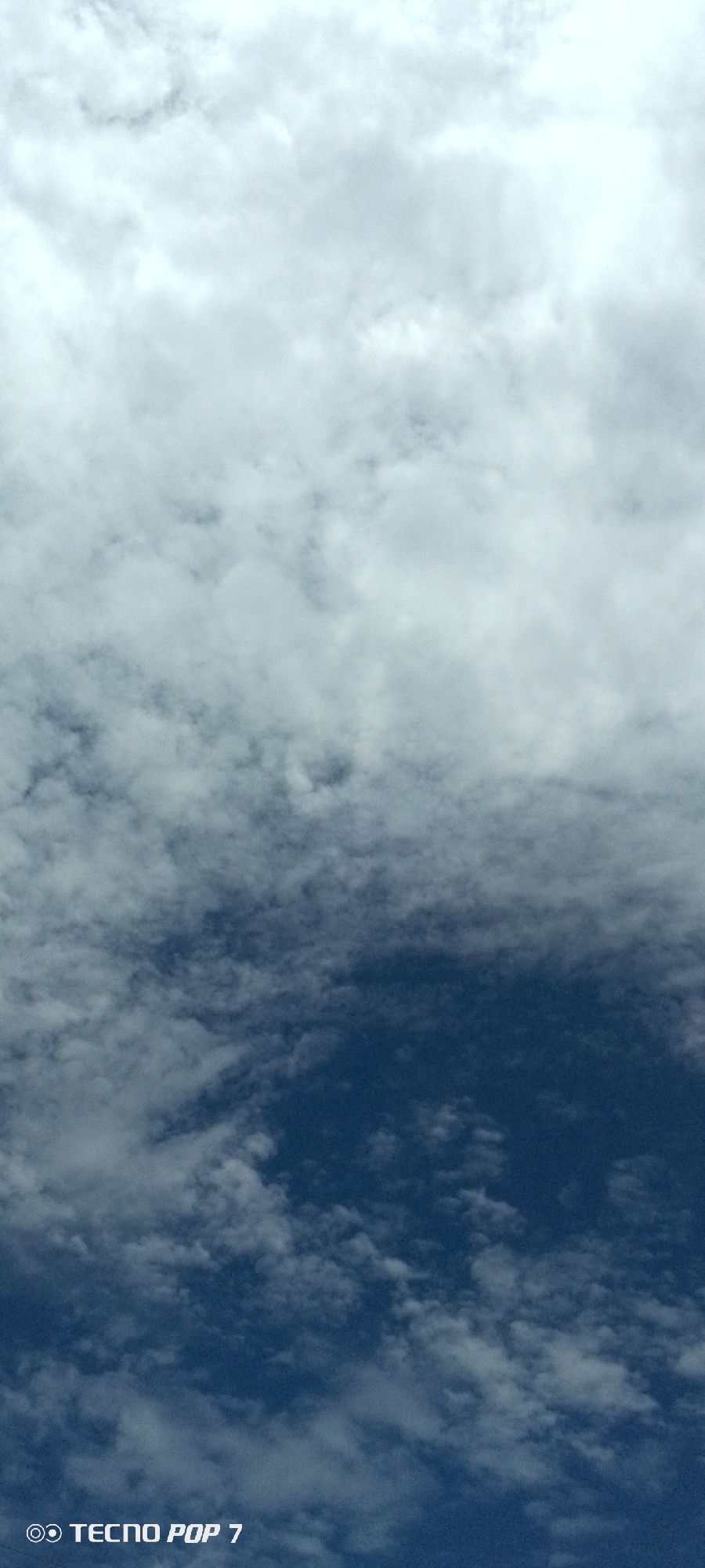 The image depicts a sky filled with clouds, transitioning from a brighter, overcast sky at the top to a deeper blue sky towards the bottom. White and gray clouds are scattered throughout, with some areas appearing more dense and others more wispy. In the lower portion of the image, a watermark reads "TECNO POP 7" in white text. The scene appears to be an outdoor shot, likely taken from the ground looking upwards. There are no people or identifiable landmarks within the frame. The general environment is that of an open sky.