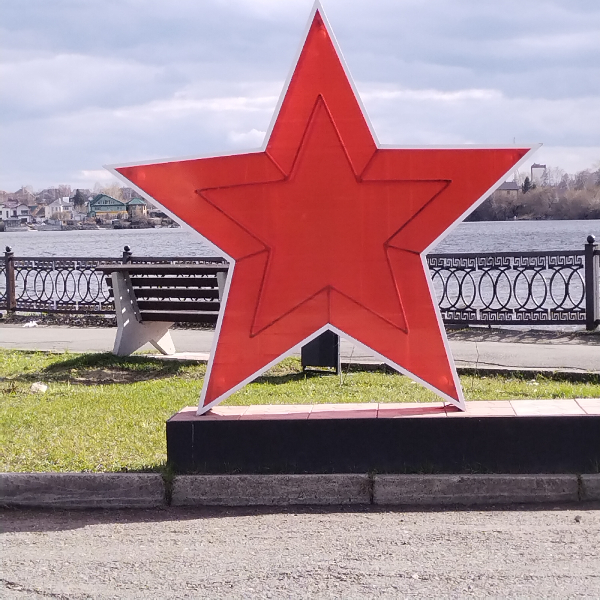 A large, red, five-pointed star, outlined in white, is prominently displayed on a tiled platform in a park-like setting. The star is situated near a paved walkway and a low concrete wall, beyond which is a grassy area with a park bench. A decorative metal fence runs along the edge of the park, separating it from a body of water, likely a river or lake, with buildings visible on the opposite shore. The sky is overcast. The location is likely in Nizhny Tagil, Russia, due to the cultural significance of the red star.