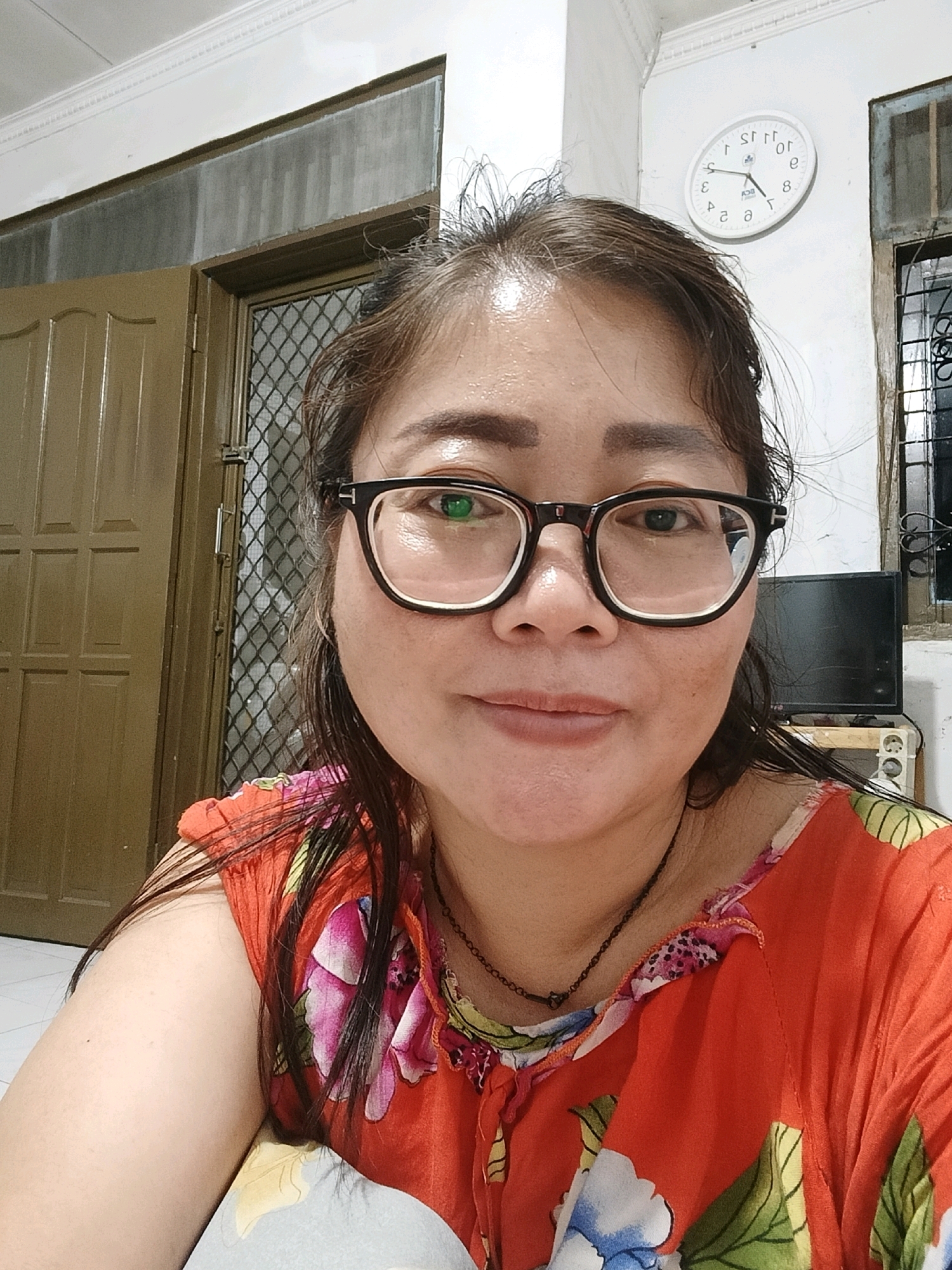 In a dimly lit room, a woman with dark hair pulled back and wearing black-rimmed glasses is captured in a close-up selfie. She has fair skin and is wearing a vibrant orange floral-patterned top. The background reveals a beige door with a diamond-patterned screen door, a white wall with molding at the top, and a white circular clock with black markings and hands mounted on the wall. To the right, a window with bars and a dark, flat-screen television on a wooden stand are visible. The environment suggests an indoor residential setting, potentially in Jatimulya, Indonesia, based on the provided context.