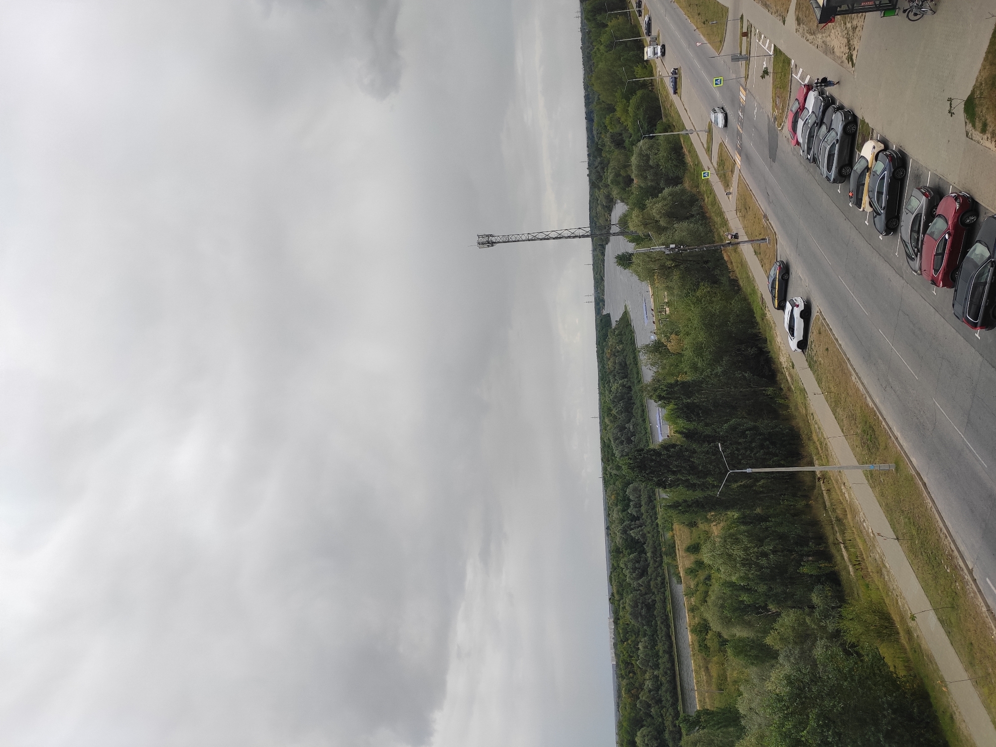 The image captures a wide-angle, high-angle view of a cloudy day in the city of Homyel, Belarus, focusing on a road with parked cars and a river with a forested bank in the background. The foreground features a paved road with several cars parked along its right side in a parallel arrangement, including a dark red SUV, a dark grey sedan, a lighter grey SUV, a silver sedan, and a white van. Further down the road, more vehicles are visible, including a white car and a larger truck. A pedestrian walkway runs alongside the road, separated by a grassy area.
Dominating the mid-ground is a wide, calm river flowing from left to right, bordered by a dense line of green trees and vegetation that stretches across the horizon. On the far side of the river, a distant line of urban buildings is faintly visible against the overcast sky. A tall, slender telecommunications tower stands prominently on the left bank of the river, overlooking the water and the surrounding greenery. The sky is filled with heavy, grey clouds, indicating an overcast or possibly rainy weather condition.