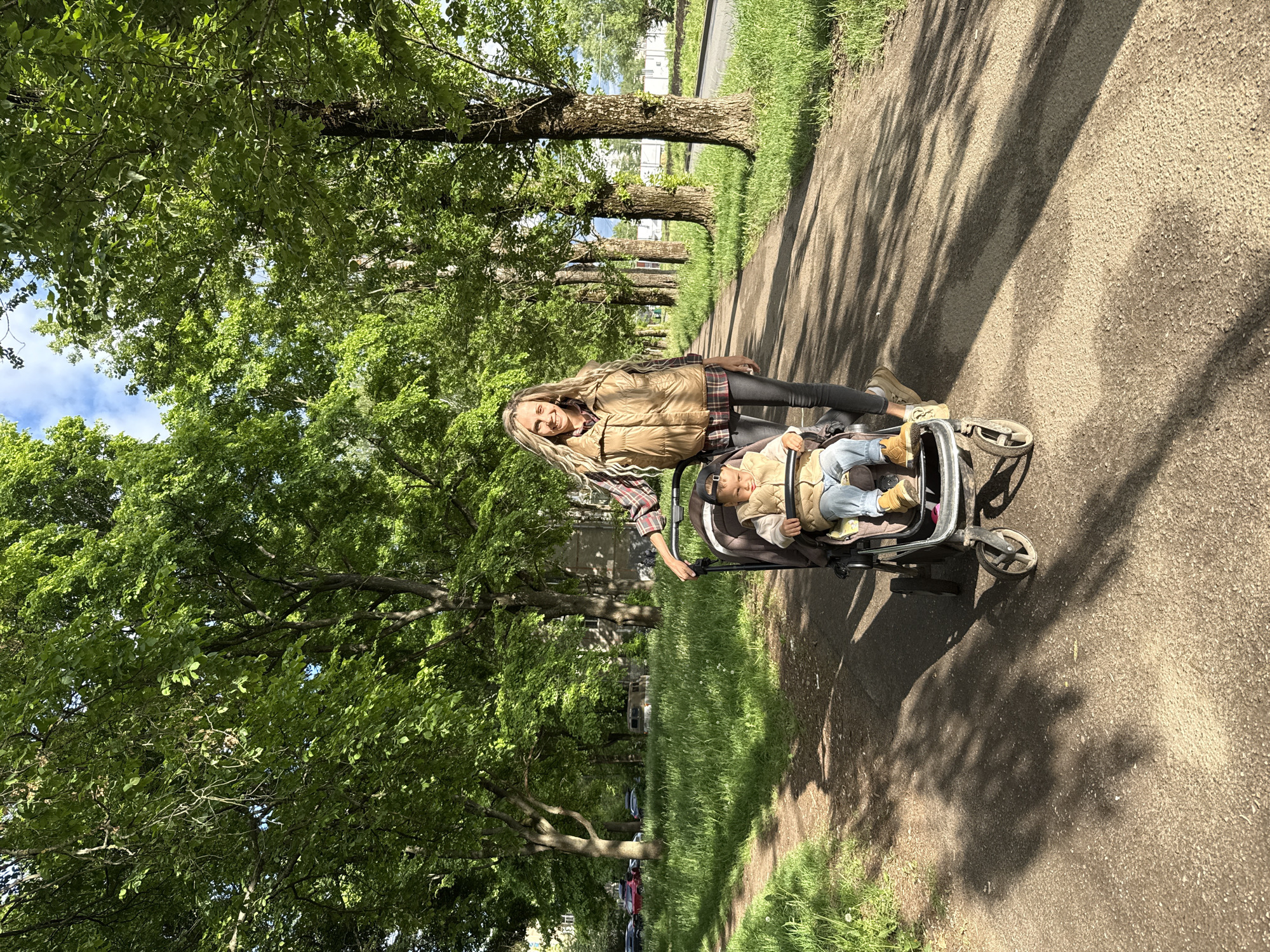 A woman with long blond hair is standing next to a baby in a stroller on a paved path lined with trees. The location is Rusanovo, Russia. The woman is wearing a tan jacket, plaid shirt, and leggings. The baby is wearing a tan jacket, jeans, and a baseball cap. The stroller is black and silver. The path is surrounded by grass and trees. The trees cast shadows on the path.