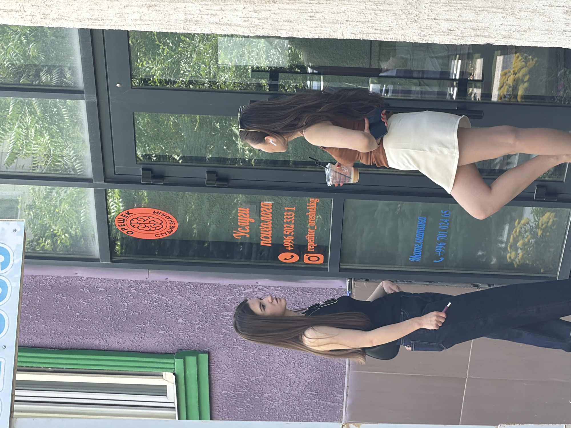 The image shows two young women standing outside a building with large windows. The woman in the foreground on the right is wearing a black top and dark jeans, holding a cigarette. She has long, dark hair. The woman in the background on the left is wearing a brown top and a white skirt, holding a drink. She also has long, dark hair. Behind them, text is visible on the window. The text includes "Услуги психолога" (Psychologist services), "+996 502 333 1", "repetitor_oreshekkg", "Математика" (Mathematics), and "+996 701 024 65". There is also a round sticker with a brain graphic and text that appears to say "ОФИ... ЗНАНИЙ". The scene appears to be outdoors, possibly in a city, during the daytime, with the sun casting some shadows. The overall mood seems casual and ordinary.