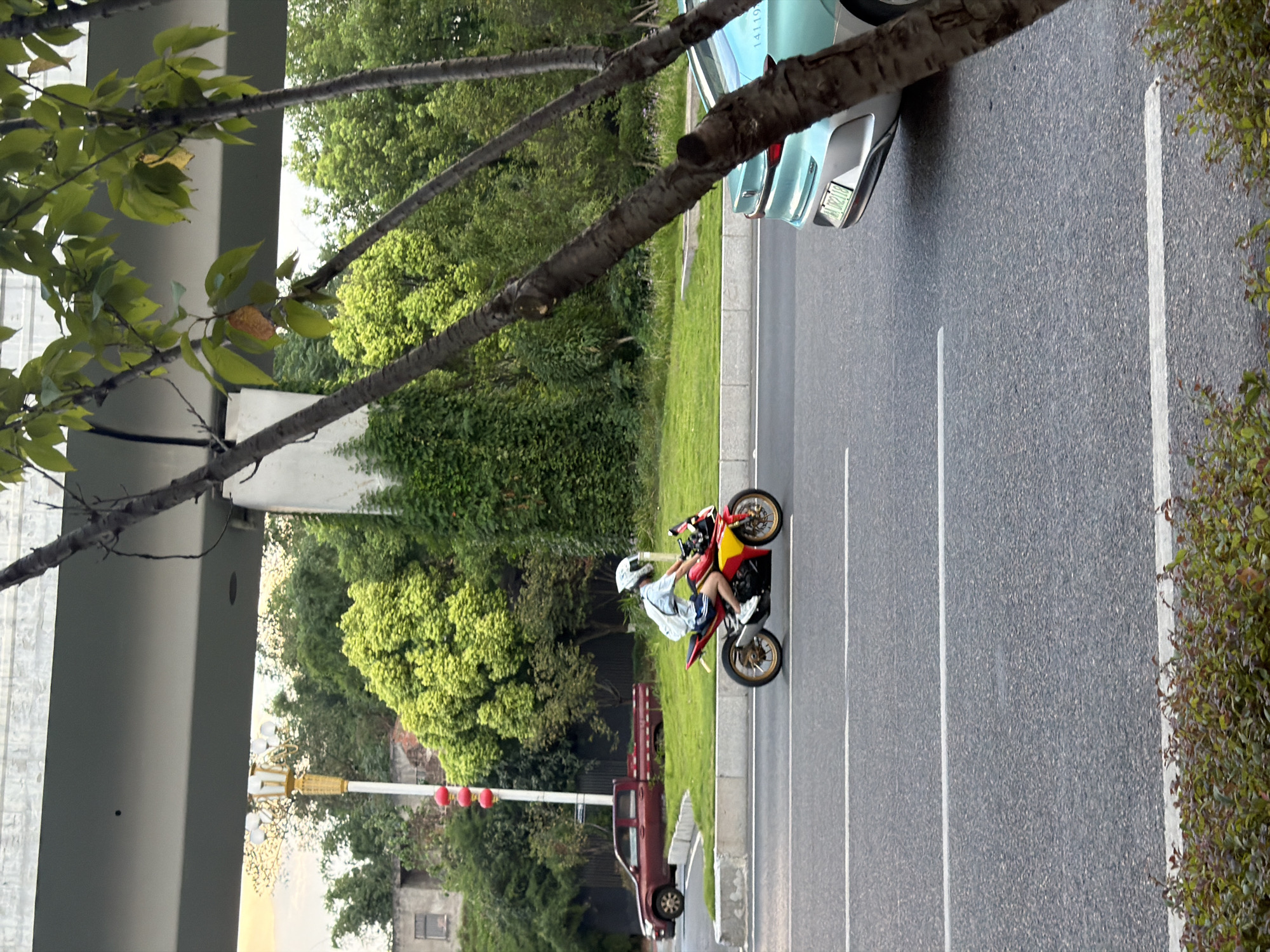 A person is riding a red and yellow motorcycle on a paved road in Jiangjin, China. The rider is wearing a white helmet, a light blue t-shirt, and dark shorts, with a white backpack. The motorcycle has gold wheels and is in motion. To the left of the motorcycle, a red pickup truck is parked. Behind the truck and motorcycle is a grassy area with trees and a building in the background. A pedestrian overpass spans across the top of the scene. In the foreground, a light blue car is partially visible on the right side of the road, and trees with green leaves are in the foreground. The road has white lane markings.