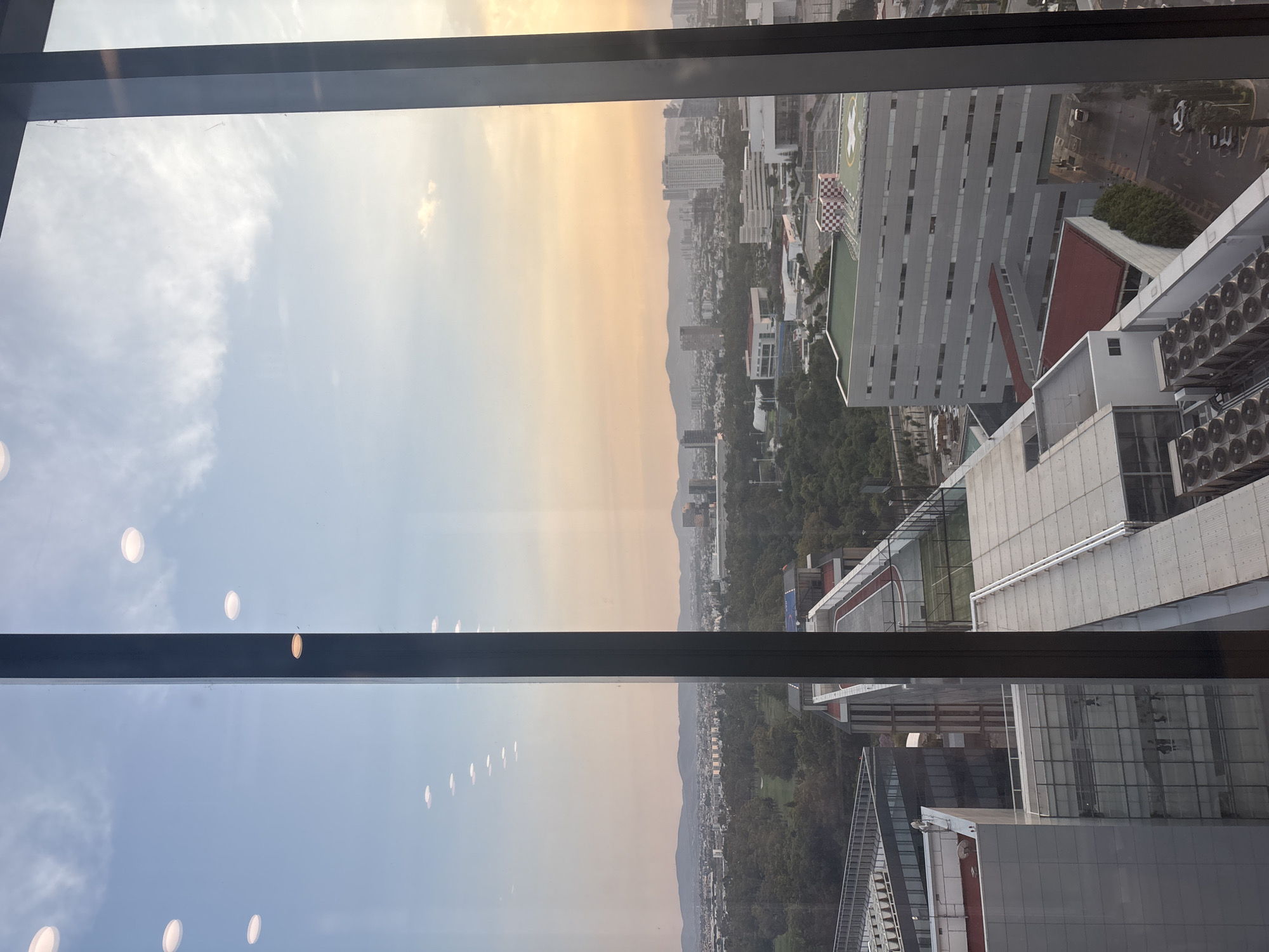 The image depicts a view from a high-rise building in Puebla, Mexico. The foreground shows parts of the building's window frame. The view overlooks a cityscape with a mix of buildings, green spaces, and distant mountains. The sky is divided with clear sky at the top left and hazy or smog-filled at the top right. Some buildings have helipads. The image appears to have been taken at dusk.