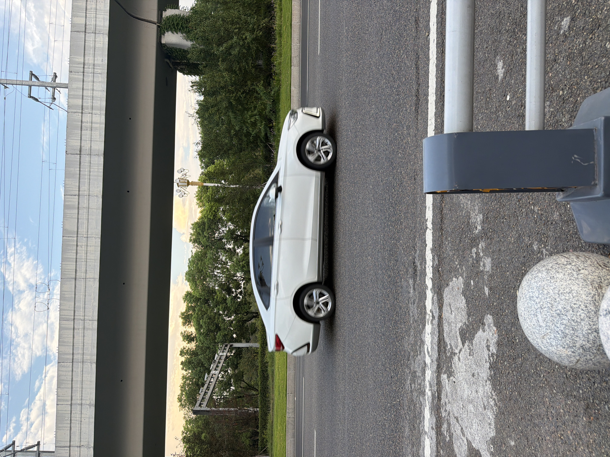The image depicts a white sedan driving on an asphalt road in Jiangjin District, Chongqing, China. The car is in motion, indicated by the blur of its wheels. In the background, a large concrete overpass spans across the scene, with visible power lines and support structures. Green trees and foliage line the side of the road, and a streetlamp with multiple lights is also present. The foreground of the image shows a portion of the road with a white line marking and a grey barrier with metal rails. Two spherical granite bollards are visible in the lower left corner. The sky above is partly cloudy, with a warm hue suggesting it might be late afternoon or early evening.