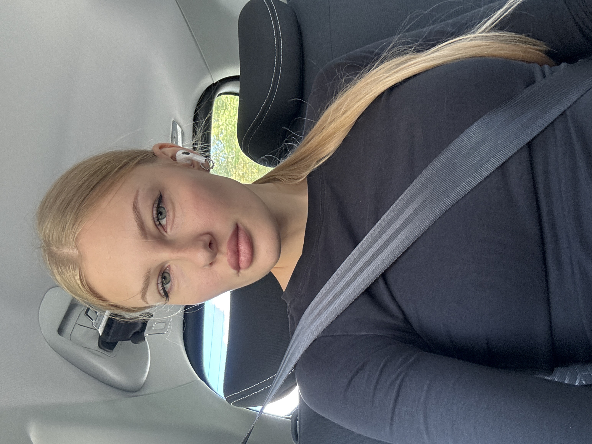 A blonde woman with green eyes and full lips is in the back seat of a car, wearing a black long-sleeved shirt and a seatbelt. She has on white wireless earbuds and a hoop earring in her left ear. Her blonde hair is pulled back into a ponytail. The interior of the car is visible, including the headrest and a part of the ceiling. Sunlight streams in from the rear window, illuminating the trees and sky outside. The photo appears to have been taken in Perm, Russia.