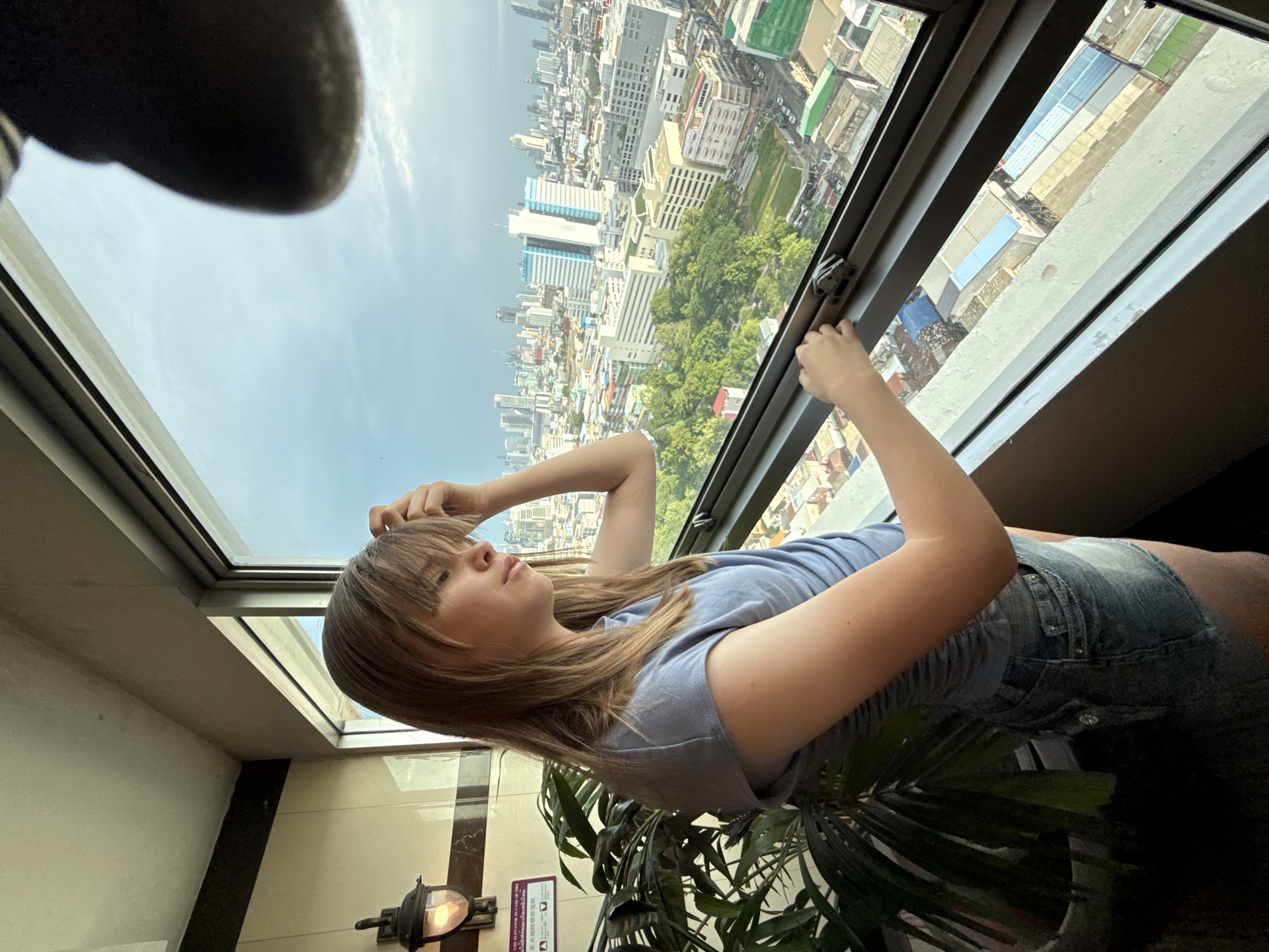 A young Caucasian female with blonde hair is standing indoors by a large window. She is looking out at the city of Bangkok, Thailand, which is visible in the background. The female is wearing a light blue t-shirt and denim shorts. She has her left hand on her head and her right hand is touching the window latch. A large potted plant is to her left, and a light fixture is mounted on the wall behind the plant. The cityscape is a dense collection of buildings, with some green spaces interspersed. The sky is partly cloudy.