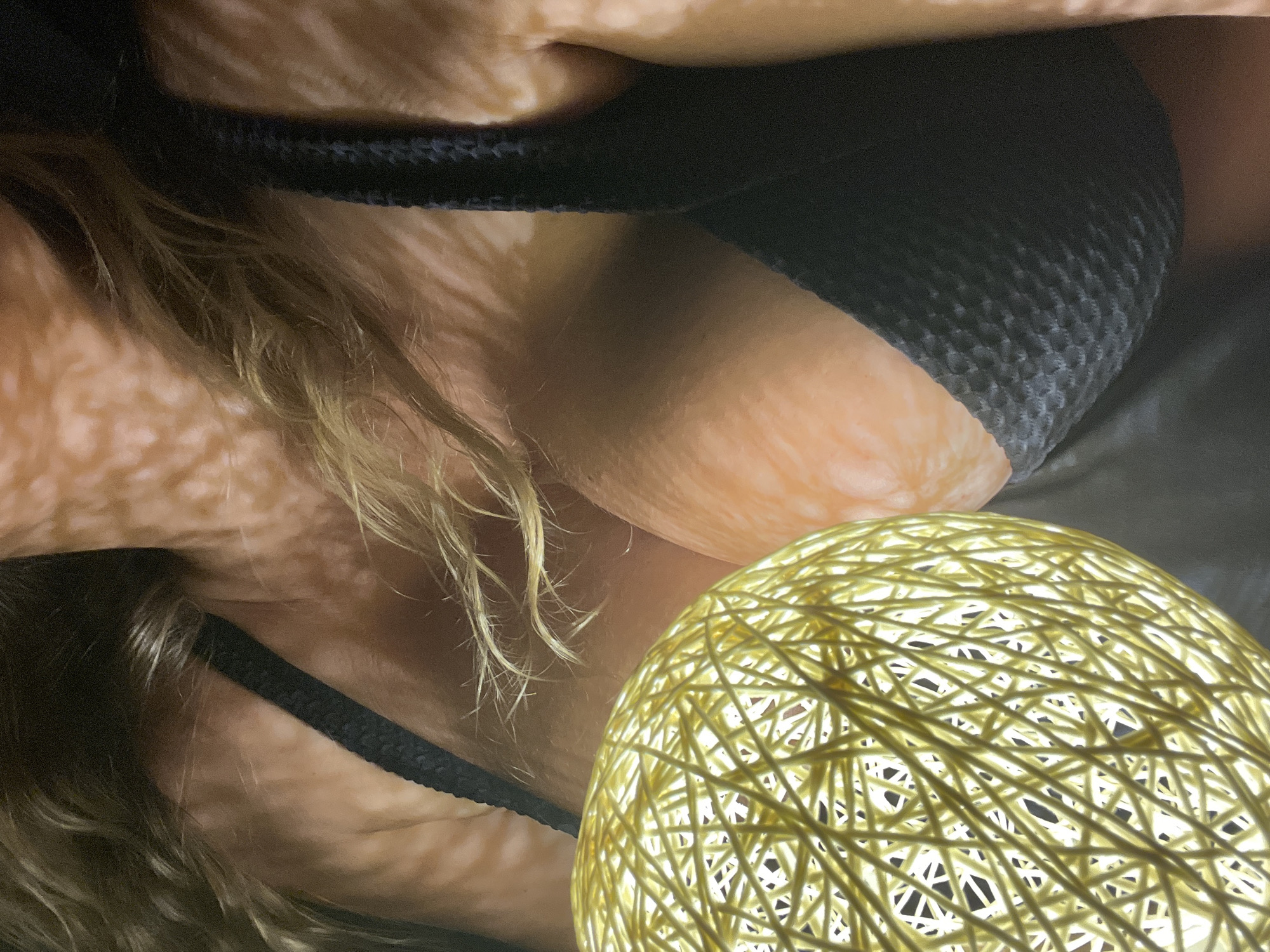 A close-up, low-angle shot shows the upper body of a person wearing a black bra. Their blonde hair is visible, framing their face which is partially obscured by a string light sphere in the foreground. The lighting creates a patterned effect on their skin.