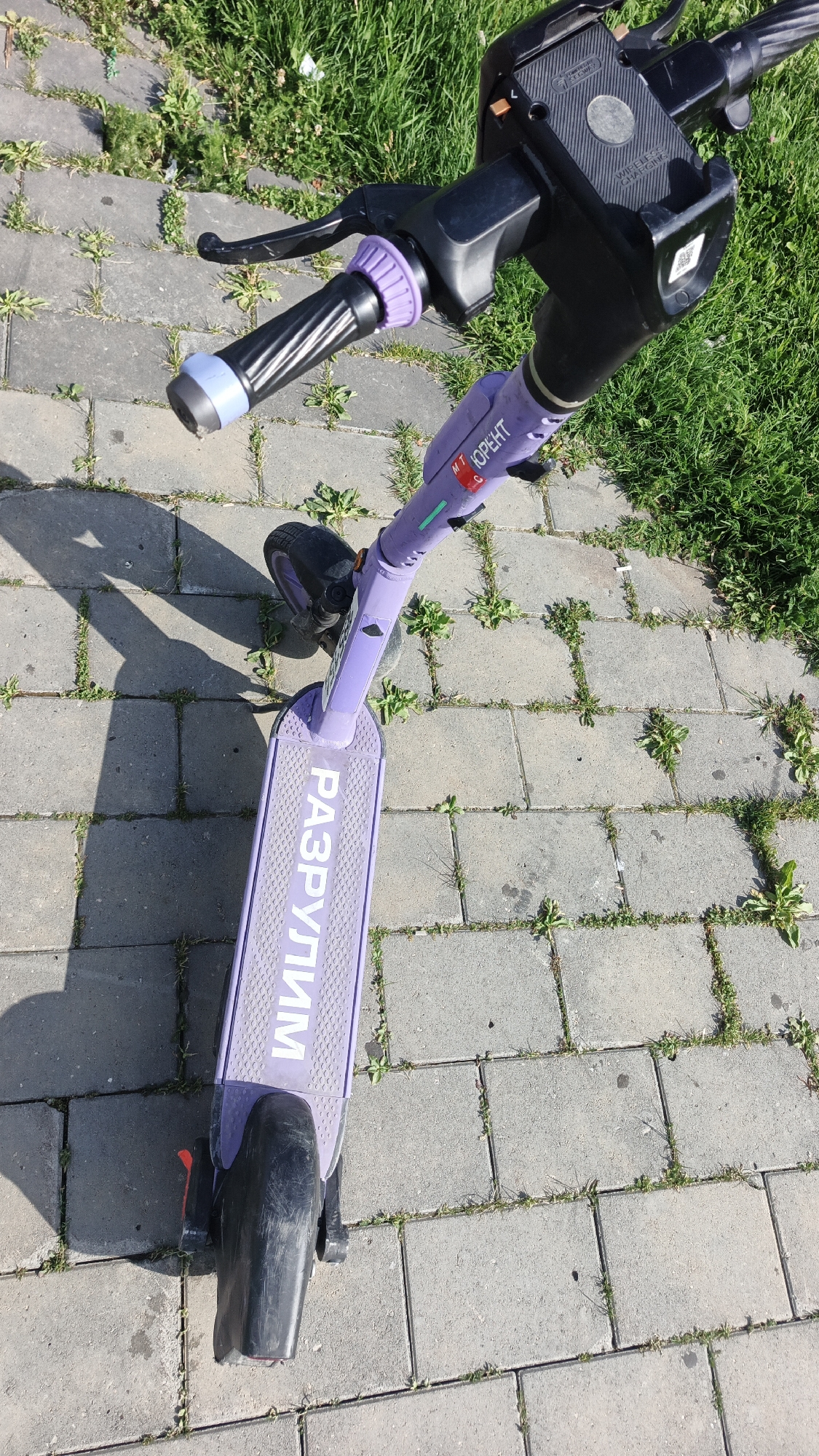 An overhead, eye-level shot shows a purple electric scooter parked on a paved walkway with grass patches. The scooter's handlebars are black with purple grips, and a small red and white sticker is visible on the steering column. The deck of the scooter is also purple and features white text that appears to read "РАЗРУЛИМ" in Cyrillic. The scooter's rear wheel and fender are black. The walkway is made of gray interlocking pavers, with green grass growing in the gaps. The background consists of more grass and a hint of sunlight casting shadows. This scene is likely located in Yekaterinburg, Russia, given the Cyrillic text on the scooter.