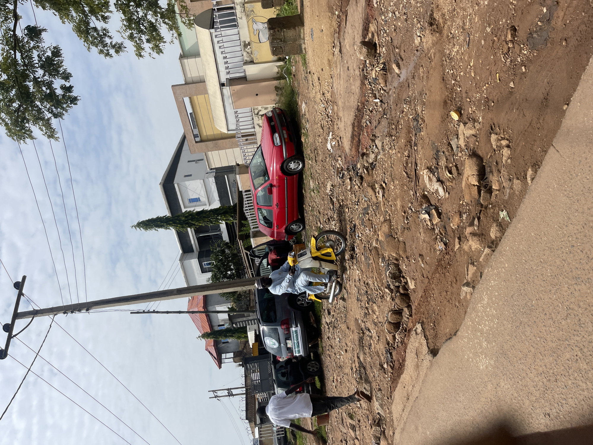 A street scene in Jos, Nigeria, features a dirt road with potholes and debris. A man in a white shirt and dark pants walks away from the camera, carrying a bucket. A second man, dressed in a light blue tunic and trousers, rides a yellow and white motorcycle. Several parked cars are visible, including a red Volkswagen Golf and a grey minivan with text on its side. In the background, modern houses with red and white roofs stand next to a large building with a beige facade and a brown accent. Overhead, power lines crisscross the sky under a cloudy, bright sky.