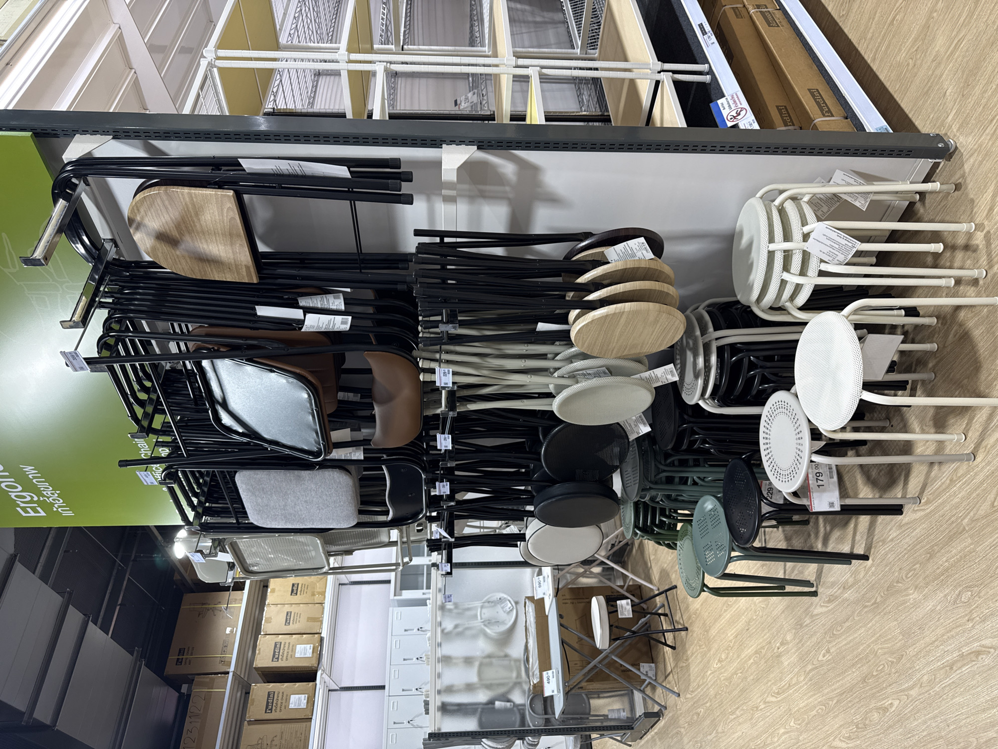 The image depicts the interior of a retail store, likely a furniture or home goods establishment, in Baan Chaweng Noi, Thailand. The scene is dominated by a display of various chairs and stools. In the foreground, several stacks of white stools with perforated seats and metal legs are visible. To the left, a row of stools in dark green and black colors, also with perforated seats, are arranged. A small folding table with a light grey surface and metal legs stands to the left of the stools.
Above the stools, a rack displays numerous folding chairs. Some chairs have black metal frames and seats of various materials, including grey fabric, black leather, and brown leather. Several chairs with light wooden seats are also present, hanging from the rack. A sign with green text, possibly an advertisement or product description, is mounted on the wall above the chairs.
In the background, shelves are lined with boxes, suggesting additional merchandise. The flooring is light-colored wood. The overall impression is of an organized retail environment showcasing seating furniture. There are no people visible in the image.