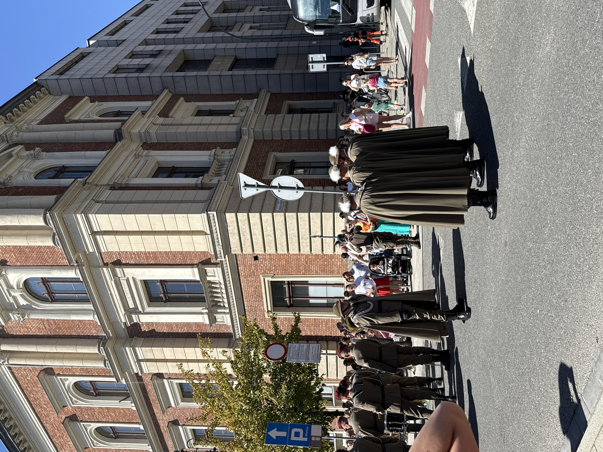 The image captures a daytime street scene in Krakow, Poland. In the foreground, a group of individuals in military-style uniforms with capes are standing on asphalt. Some are wearing dark boots and hats, and one is holding a rifle. Behind them, a crowd of onlookers, including men, women, and children, are observing. Some are holding cameras, and one person is pushing a stroller.
The background features two buildings: a historic red brick building with classical architectural elements and a more modern, grey, multi-story building. There are trees and street signs visible, including a blue sign with a white arrow and the letter "P," indicating parking. A bus is partially visible on the right side of the frame. The sky is clear and blue. The scene appears to be part of a public event or parade.