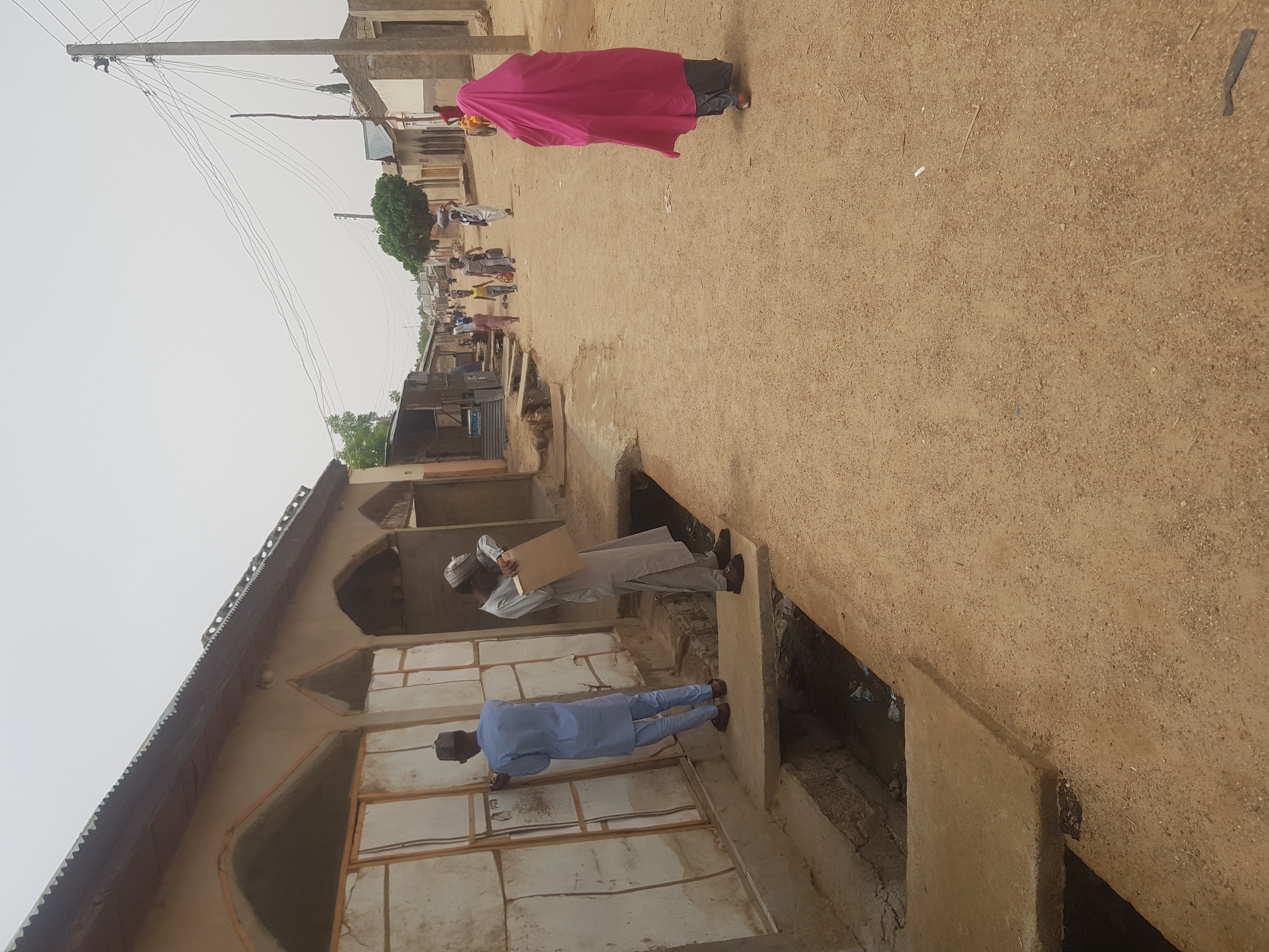 In Dutsinma B, Nigeria, the scene depicts a partially dirt road in a built-up area. A woman in a pink covering walks away from the camera. Several other people are visible in the distance. A man wearing a blue garment stands in the foreground, while another man wearing a gray garment stands next to him and holds a cardboard item. They both appear to be near a building with arched entryways. The street is unpaved, and a drainage system is visible along the edge. There are utility poles and wires visible in the sky.