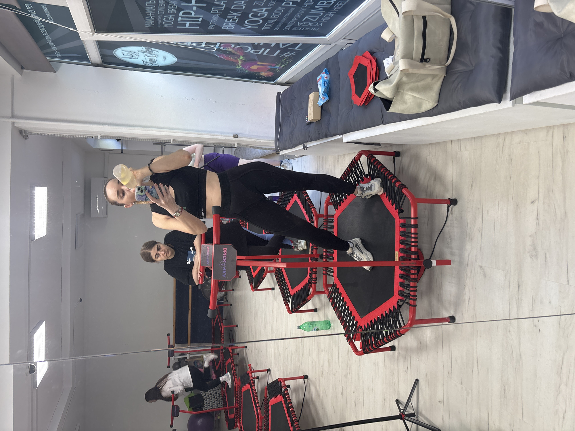 The image shows a gym with multiple rebounders, which are small trampolines used for exercise. In the foreground, a woman is captured in a mirror reflection, holding a water bottle and her phone, seemingly taking a selfie while exercising. She is wearing black athletic wear. Behind her, another woman in purple athletic wear is also on a rebounder. The gym appears to be well-lit, suggesting it's daytime or under bright artificial lights. A gray mat with red hexagon-shaped mats is visible on a raised platform, and a beige backpack is placed on it. The floor is light-colored wood laminate. Visible text on the wall includes "HIP-H", "YOGA BREAK", "CONTEMPORARY HIP", "ZUMBA", and "#ПРОС". The overall scene suggests a fitness class or individual workout session is in progress.