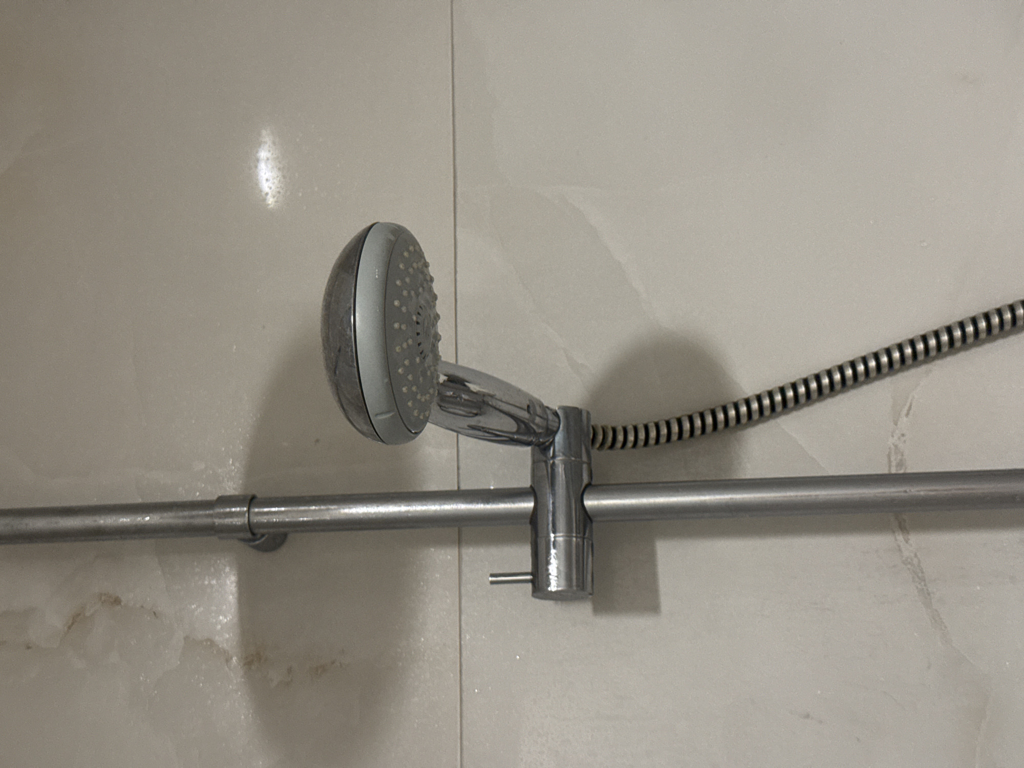 A close-up, eye-level shot shows a chrome shower head on a vertical riser rail against a pale tiled wall. The shower head is oval-shaped with visible spray nozzles and is attached to the rail by a flexible, ribbed hose. The rail itself is secured to the wall by a bracket. The wall tiles have a slight marbling effect, and there are faint water stains and residue visible. The lighting is even, suggesting an indoor environment, possibly a bathroom in Aksu, Turkey. There are no people or animals in the frame.