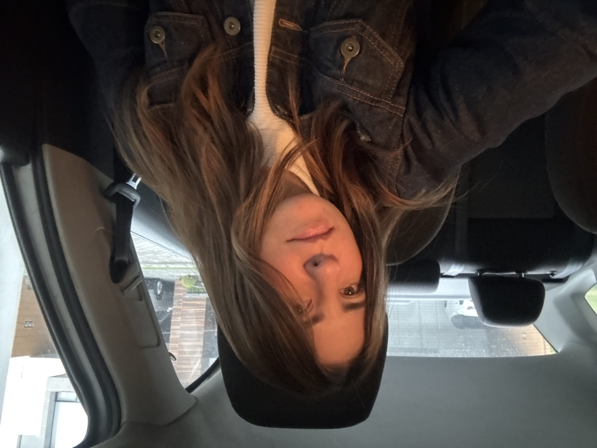 A female with light brown hair and brown eyes is sitting in the back seat of a car. She is wearing a white top and a dark denim jacket. The seat is black leather. Outside the window, a glimpse of the city shows a rainy street, suggesting a cloudy day. The location is likely in Puebla, Mexico, assuming that information is pertinent to the context.