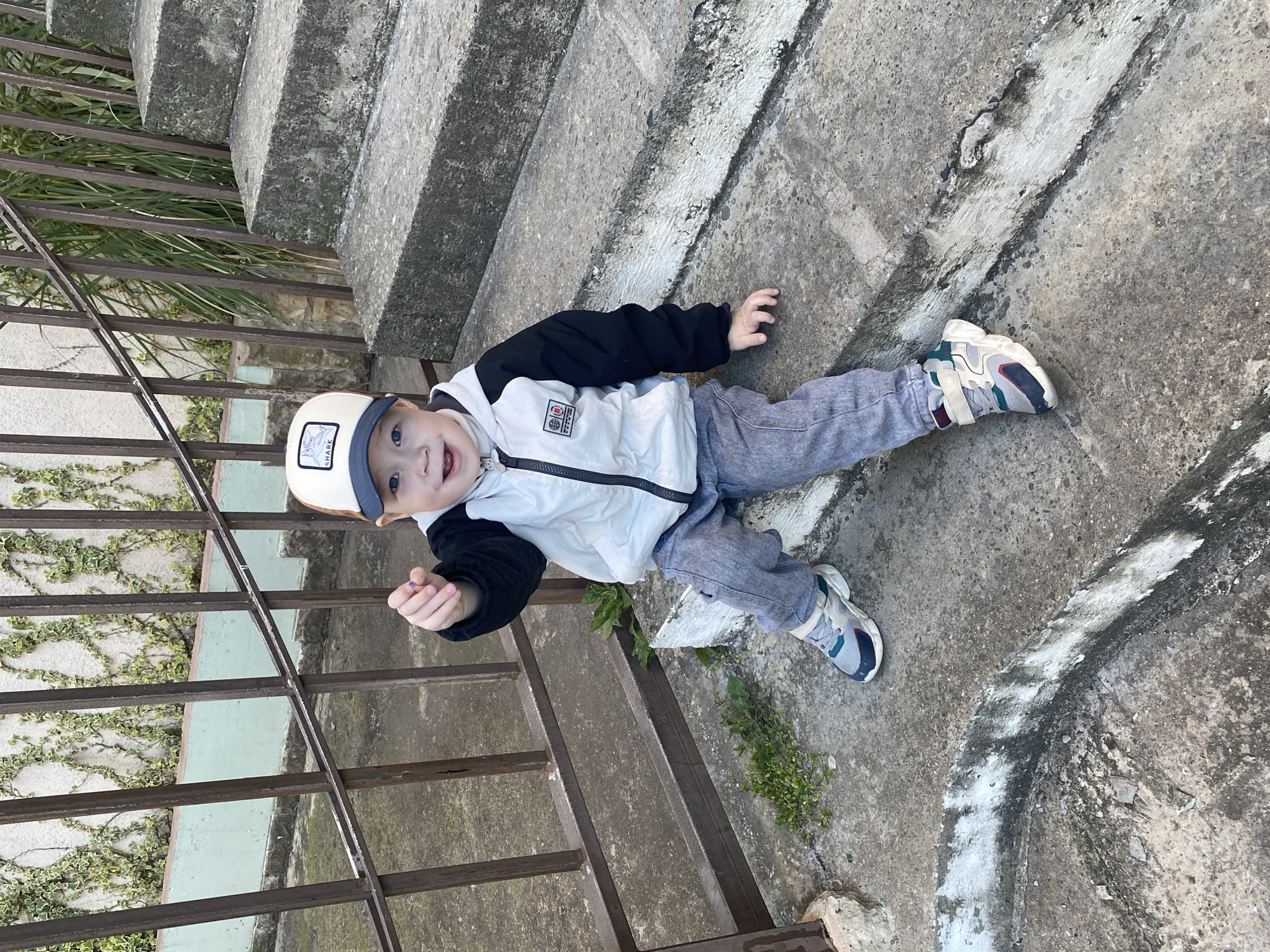 The image, taken in Sochi, Russia, features a young child, likely a boy, lying on a weathered concrete surface next to a set of concrete steps and a metal railing. The child is smiling broadly and looking directly at the camera, conveying a sense of playfulness and happiness.
He is dressed in a two-toned jacket with a white body and black sleeves, grey trousers, and colorful sneakers predominantly in white, blue, and grey with hints of red. A cream-colored baseball cap with a blue brim and a patch featuring a shark motif and the word "SHARK" is worn on his head. In his left hand, the child holds a small, indistinct purple object.
The setting is an outdoor area, likely a public or utilitarian space, characterized by robust concrete steps and a sturdy metal railing that runs parallel to them. Behind the railing, green grass or tall weeds grow near the steps, and further left, ivy climbs a textured wall. The concrete ground shows signs of wear, with patches of white and dark discoloration, and small green plants sprout from cracks. The lighting suggests it is daytime, with bright, natural light.