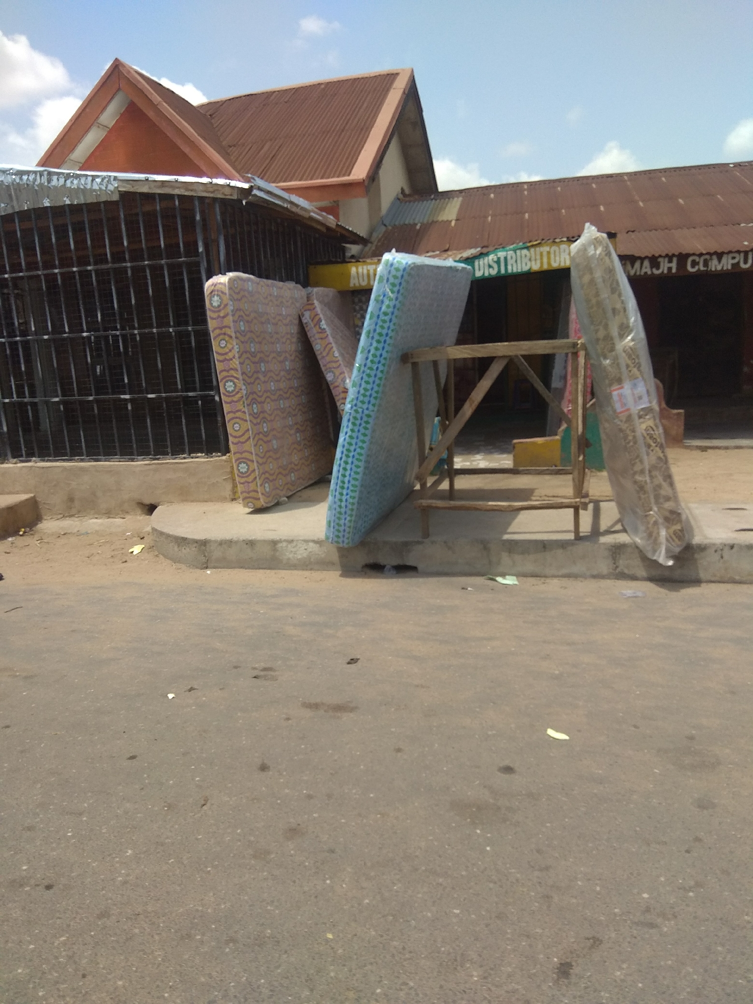 The image depicts an outdoor scene in Jalingo, Nigeria, likely a commercial area. In the foreground, a paved road is visible, with some debris scattered on it. A concrete curb separates the road from a sandy area, which then leads to a building.
Several mattresses are displayed in front of the building. One mattress, with a patterned cover featuring orange, purple, and white designs, leans against a wooden stand. Another mattress, covered in a blue and green checkered pattern, is propped up behind the first. A third mattress, wrapped in clear plastic and bearing a label, stands tall on the right side of the wooden structure. The wooden stand itself appears rustic and homemade.
Behind the mattresses, the building has a corrugated metal roof, some parts of which are rusted. The building's facade features a section with a reddish-orange sloped roof and barred windows. Above the windows, there is a sign with green lettering that reads "DISTRIBUTOR" and "MAJH COMPU". The time of day appears to be daytime, with clear, bright lighting suggesting fair weather. The overall impression is one of a retail space, possibly a mattress shop or a general store, operating in a tropical or sub-tropical environment. There are no people visible in the image.