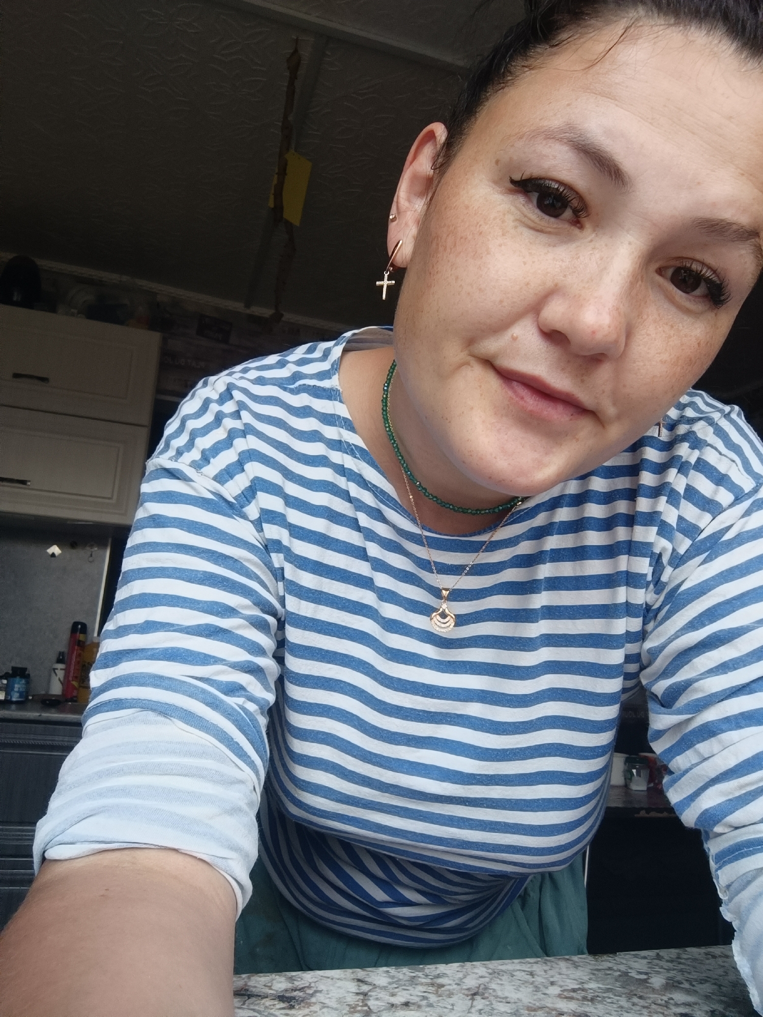 A woman with brown hair pulled back is shown in a selfie. She has light skin with freckles and dark brown eyes. She is wearing a blue and white striped long-sleeved shirt and a green necklace with a gold pendant. A gold cross earring is visible in her left ear. She is leaning forward and looking at the camera. In the background, a kitchen area is visible with cabinets and countertops. The scene appears to be indoors. There is no information in the image to suggest the location is the city of Karsha, Russia.