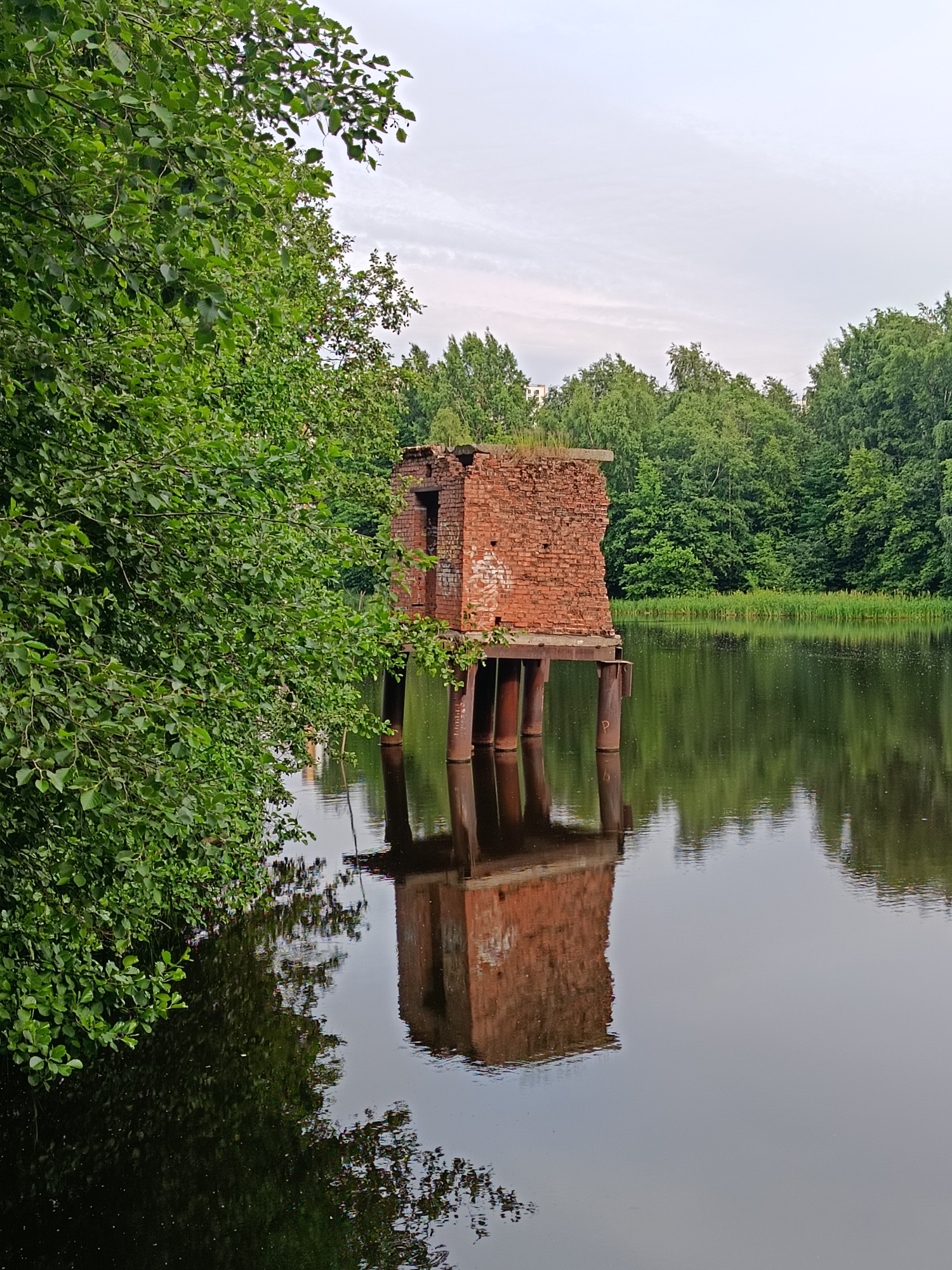 An abandoned brick structure, likely a former observation or defensive post, stands on stilts in a dark body of water. The structure is rectangular with a partially collapsed roof and an opening on one side. It is supported by cylindrical metal pilings. Lush green trees and foliage surround the water, with the dense forest extending to the horizon. The sky above is overcast and light gray. The water reflects the structure and the surrounding trees, creating a symmetrical image. The scene evokes a sense of decay and nature reclaiming man-made structures. The location is identified as the Fort "Krasnaya Gorka" in the city of Saint Petersburg, Russia.