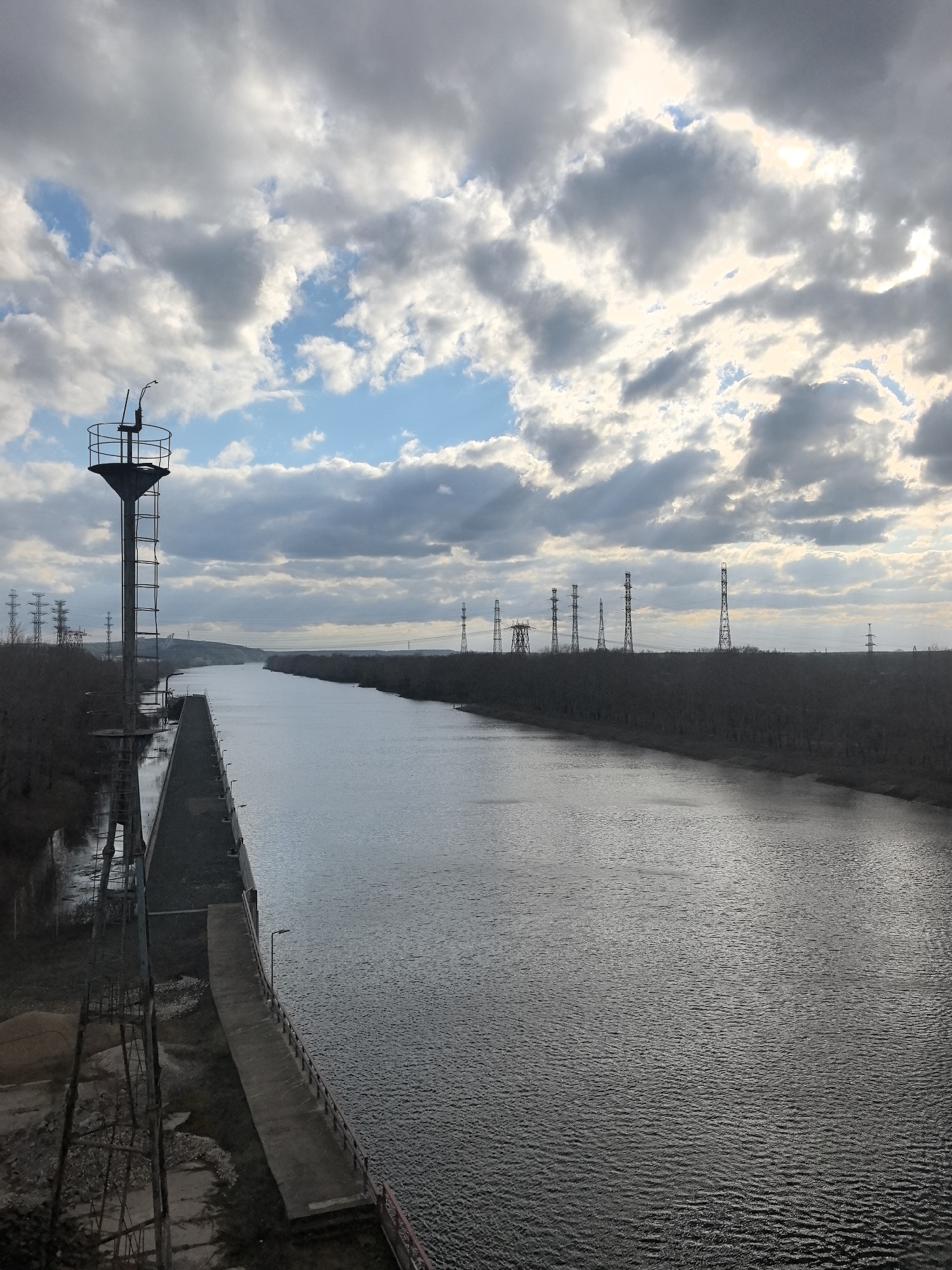 The image presents an expansive view of a wide, straight canal or river, likely the Kama River in Chaikovsky, Russia, under a dramatic, cloudy sky. The water surface, textured with numerous small ripples, reflects the muted daylight.
On the left bank, a tall, slender metal tower dominates the foreground. It features a circular platform at its apex, complete with a protective railing and what appears to be a small antenna or light fixture. A vertical ladder provides access up the tower's shaft. Adjacent to the tower, a long, dark concrete embankment or pier stretches along the river's edge, leading to a small concrete landing with a lone lamppost. The immediate bank area around the tower is rugged, showing traces of earth and sparse, bare vegetation.
Both banks of the waterway are lined with dense, deciduous trees or bushes, appearing leafless, which suggests the season is late autumn or winter. Along the distant right bank, a series of high-voltage electricity transmission towers, or pylons, are silhouetted against the horizon, marking an industrial presence within the landscape. Additional pylons are visible further in the background on the left side as well.
The sky is a prominent feature, covered mostly by a dynamic array of grey and white clouds, through which several patches of bright blue sky are visible. The lighting is diffused, indicating daytime, possibly late afternoon, with a soft glow emanating from behind the clouds. There are no people or active events visible in the scene, which evokes a sense of tranquil, industrial scenery integrated with nature. No visible text is present in the image.