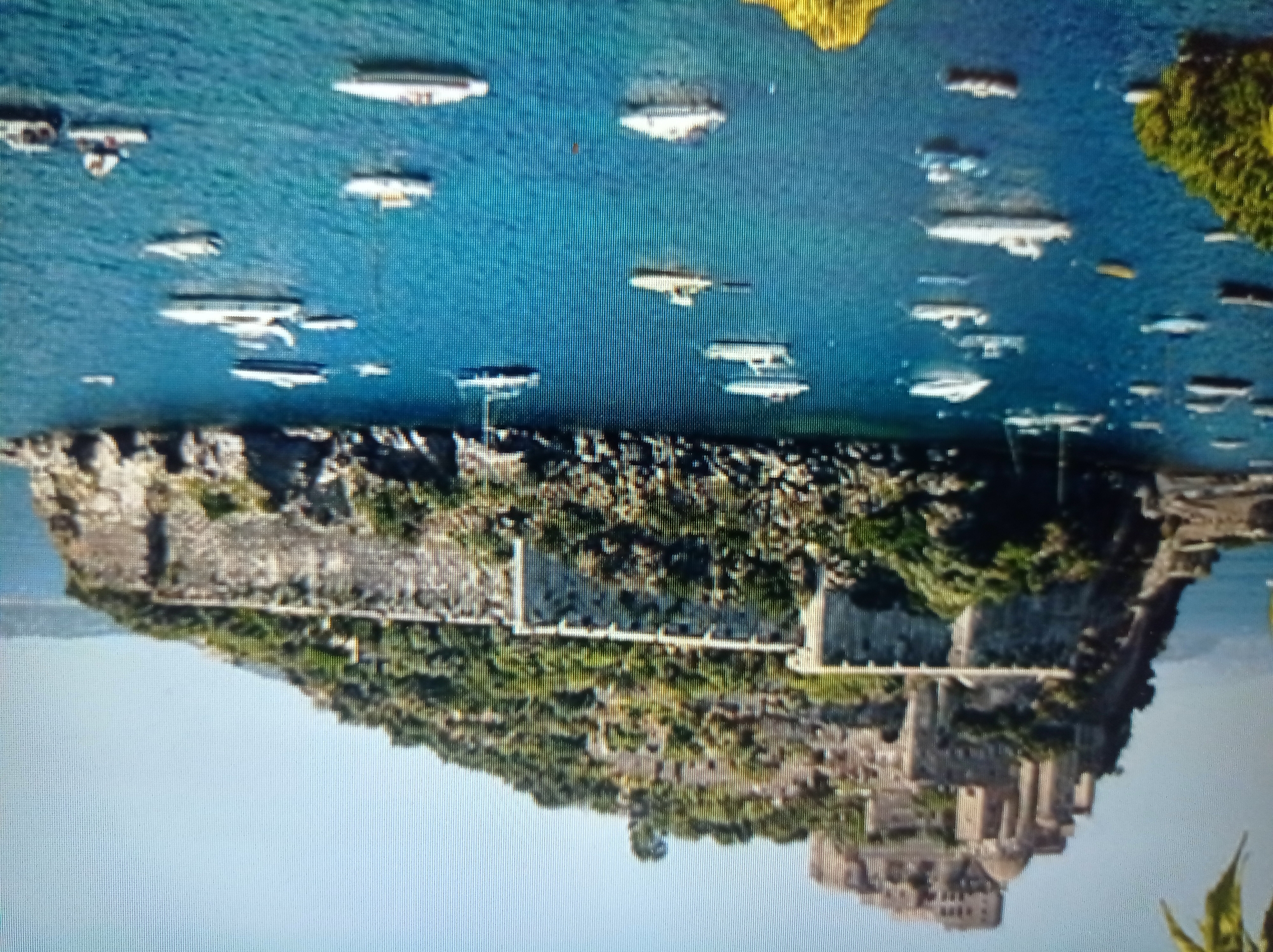 The image depicts a historical castle perched atop a lush, green island, overlooking a body of bright blue water dotted with numerous boats of various sizes. The castle complex features stone walls, towers, and buildings, indicating a significant historical structure. The island itself is densely vegetated, with trees and shrubs covering its slopes. The surrounding water is calm and reflects the clear blue sky. The boats, mostly modern yachts and smaller vessels, are moored or sailing in the bay. The scene evokes a sense of a picturesque coastal location.
This image is not of Mukachevo, Ukraine. Mukachevo is a city located inland in western Ukraine, known for its historical Palanok Castle. The scenery in the provided image, with its coastal setting and island castle, does not match the geographical characteristics of Mukachevo.
