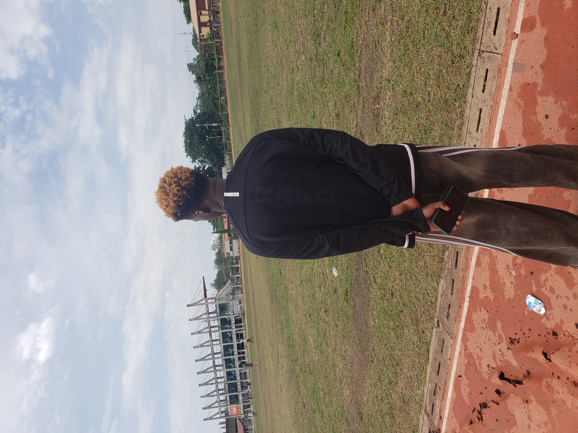 A person with bleached blonde, curly hair stands on a running track with a green field behind them. They are wearing a black long-sleeved shirt with white stripes down the sleeves and dark pants. The person is holding a smartphone in their right hand. The scene appears to be a sports field, possibly a stadium, with stadium seating visible in the background. The sky is partly cloudy. The image is taken from behind the subject, looking outwards towards the field. The ground of the running track is reddish-brown, and there are concrete dividers separating it from the grass. Some litter, including a plastic bottle, is visible on the track. The time of day appears to be daytime, with ample natural light. The weather seems mild, with the sky indicating fair conditions. The context suggests an outdoor setting in Lagos, Nigeria. There is visible text on the back of the person's shirt, appearing to be a word or logo in a dark, block font.
